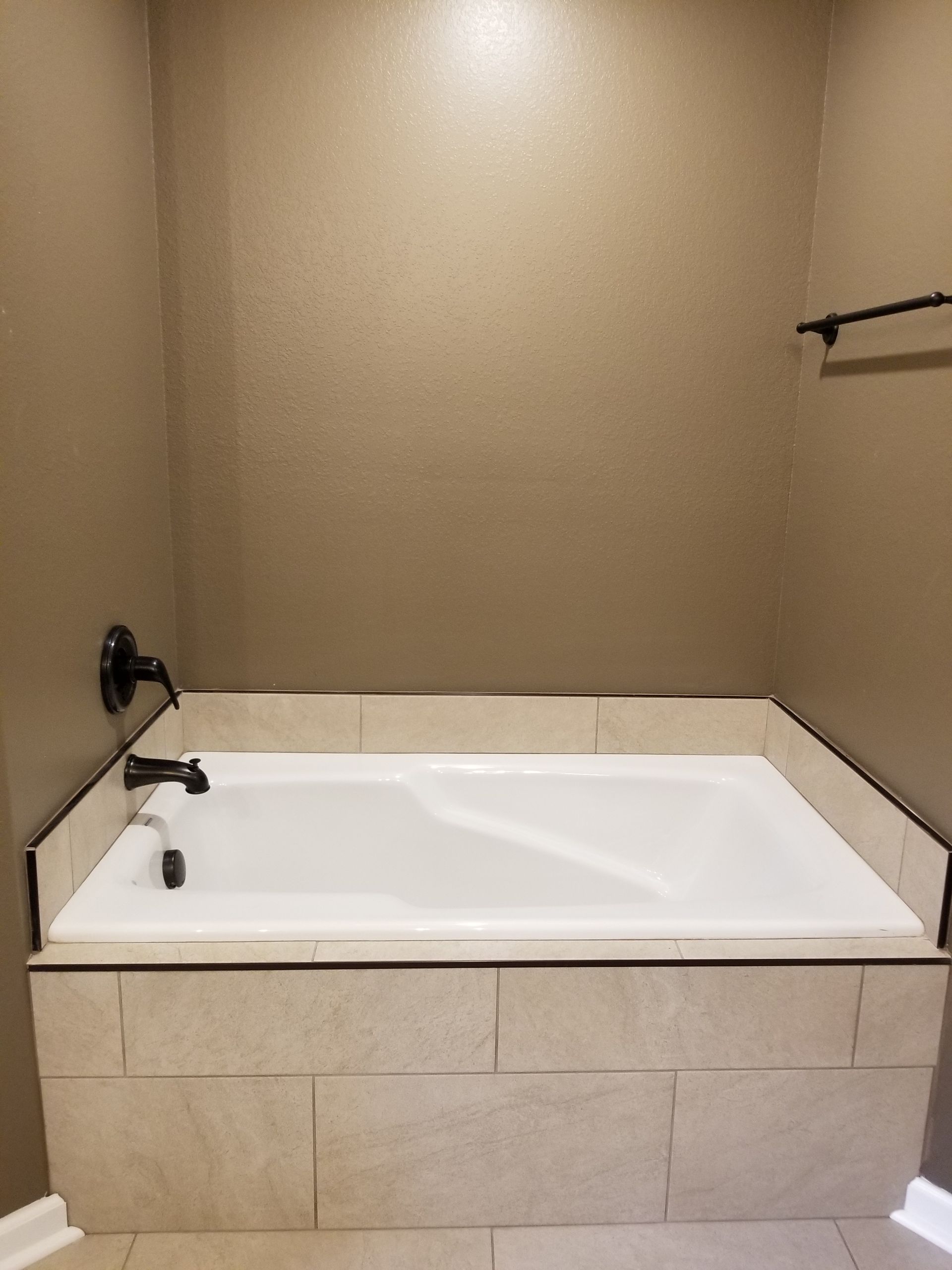 Bathtub alcove with tan tile, matte black fixtures, and a towel bar; light tan walls.