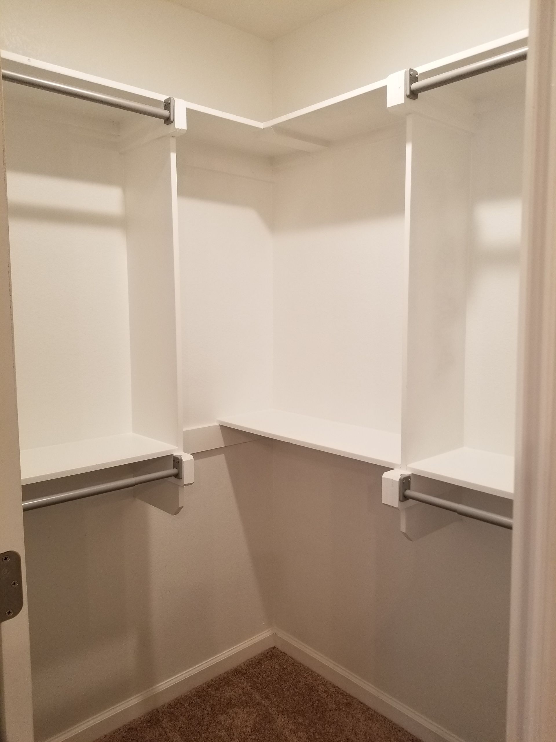 Empty, white-shelved closet with clothes rods, built into a corner, beige carpet.