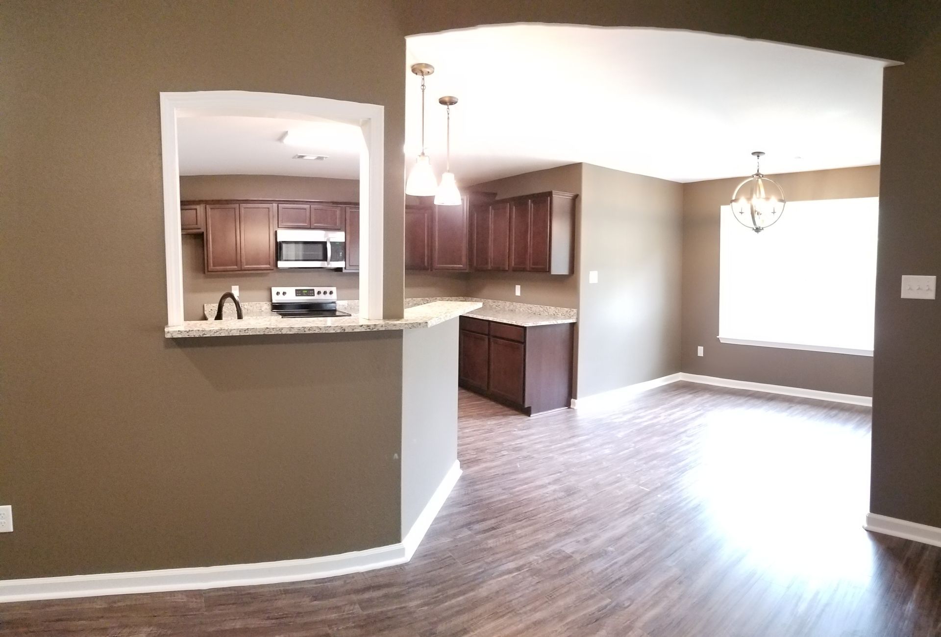 A brown-walled room with a view of a kitchen and dining area with wood floors and dark cabinets.