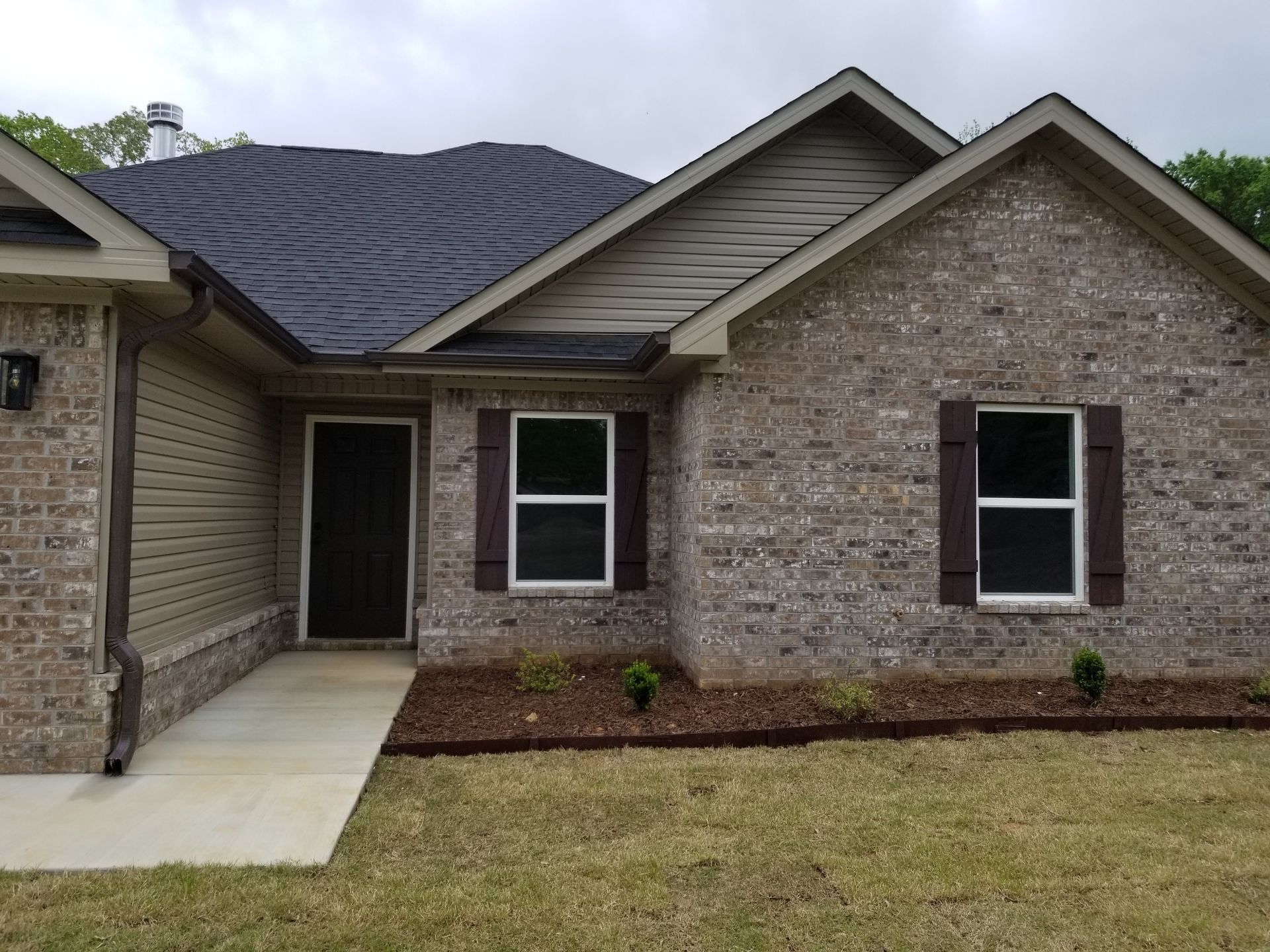 Brick house with dark roof and shutters, light siding, and a concrete walkway on a grassy lawn.