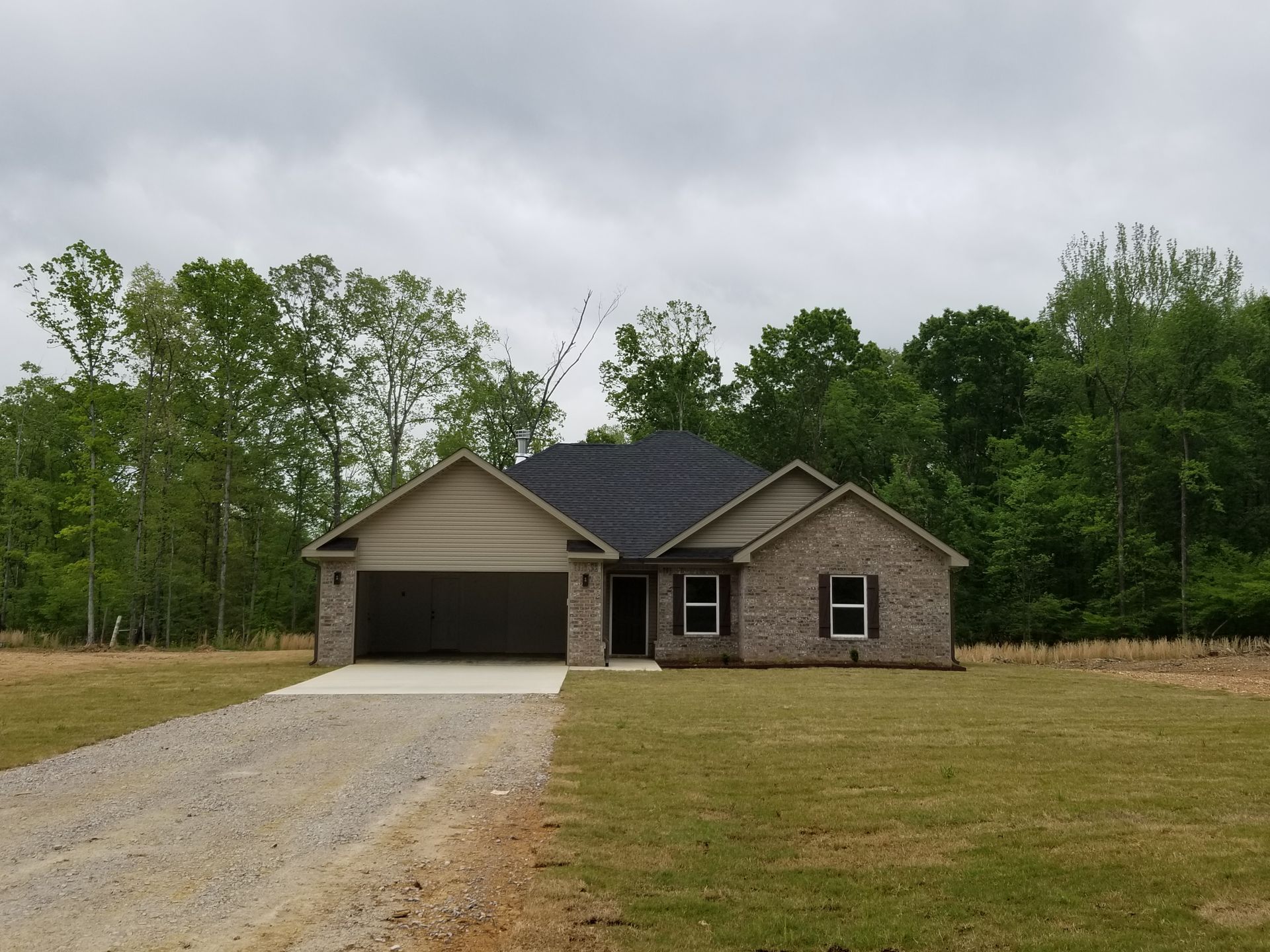 Brick house with attached garage, gravel driveway, and grassy yard; set against a backdrop of trees under a cloudy sky.