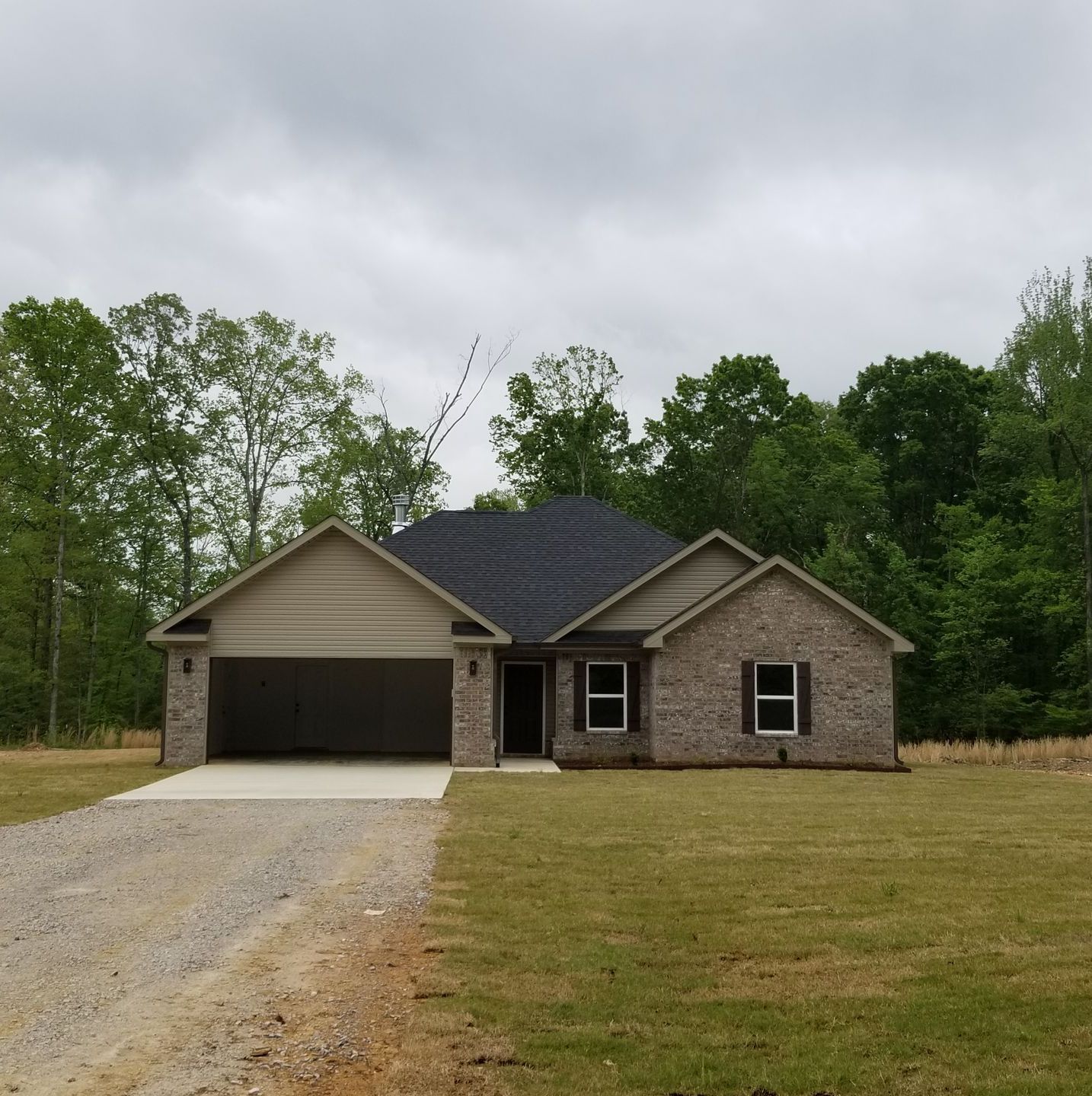 Ranch-style house with brick exterior, attached garage, gravel driveway, and green lawn under a cloudy sky.
