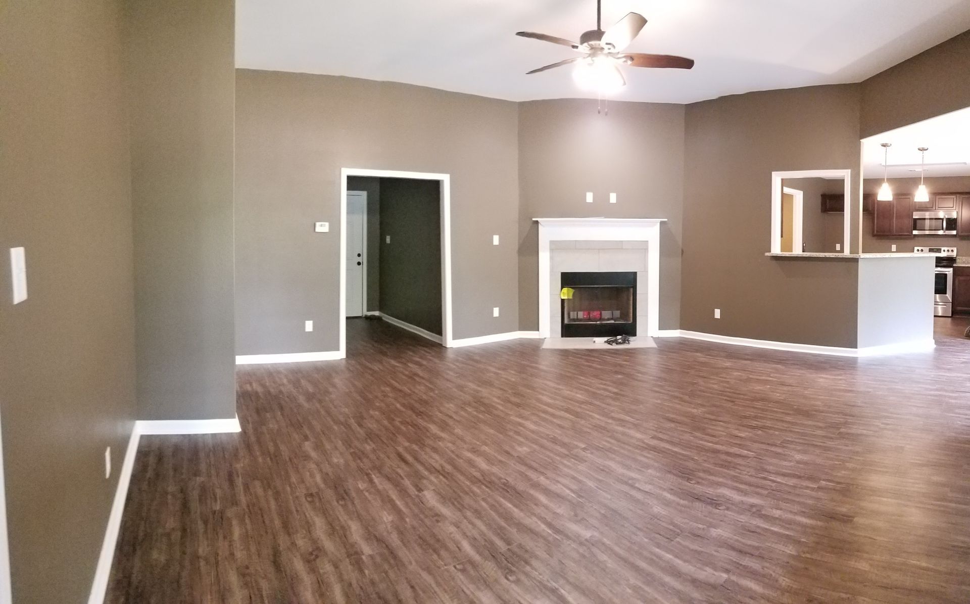 Empty living room with brown walls, wooden floor, fireplace, and ceiling fan.