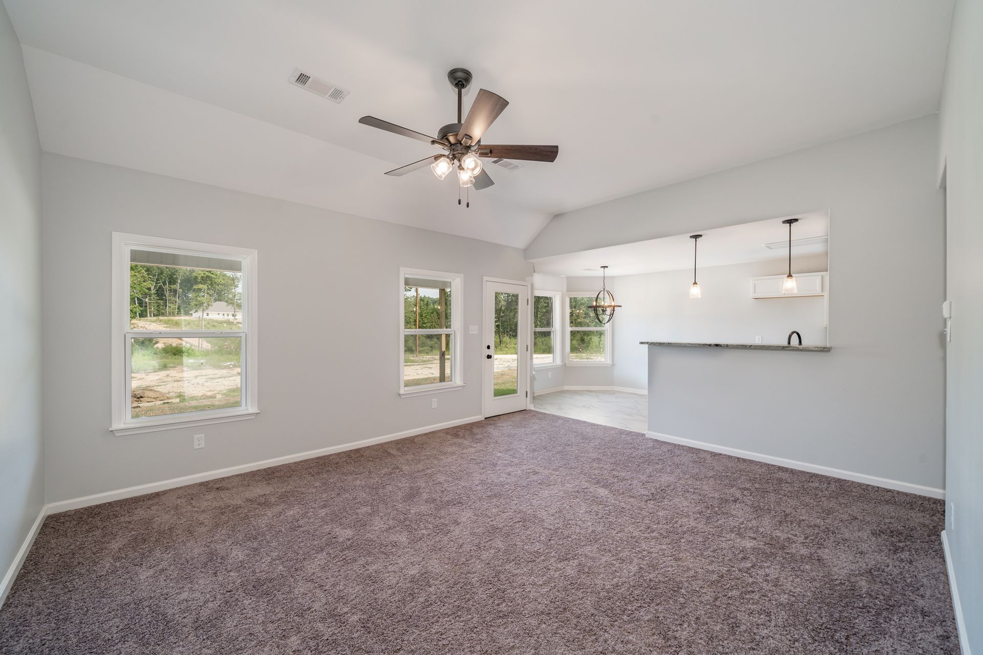 Empty living room with gray walls, brown carpet, and an open kitchen.