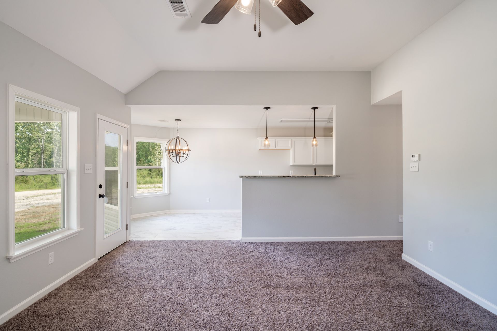 Empty dining room with gray walls, brown carpet, and partial view of kitchen.