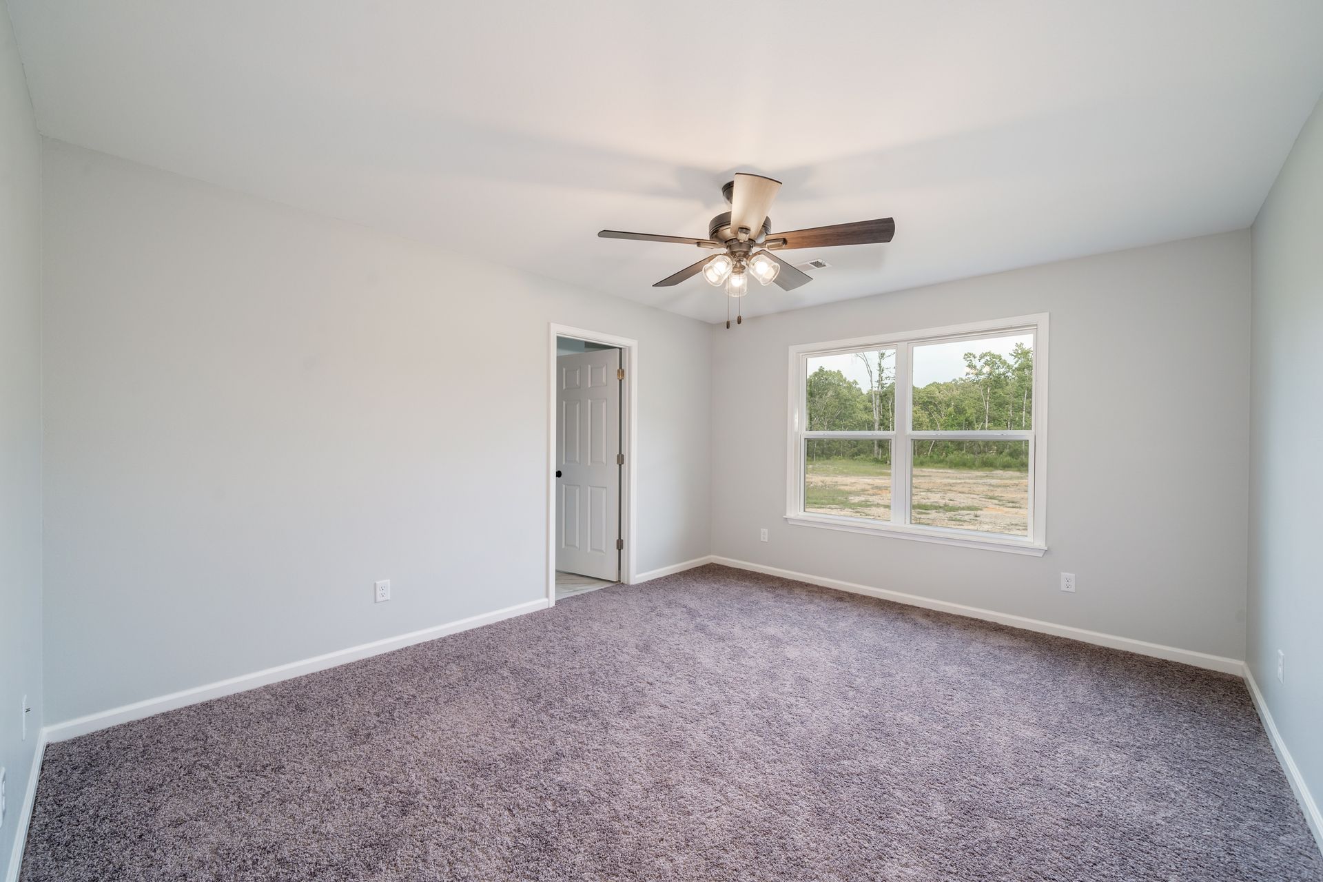 Empty bedroom with light gray walls, dark brown carpet, window, and ceiling fan.
