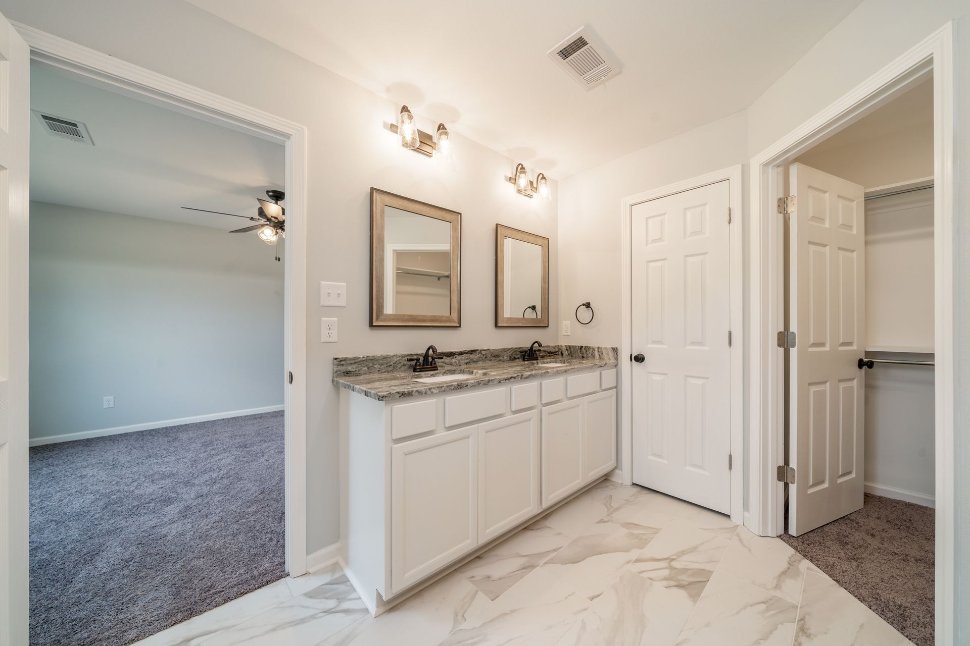 Bathroom with a double vanity, mirrors, and a closet; a bedroom is visible in the background.