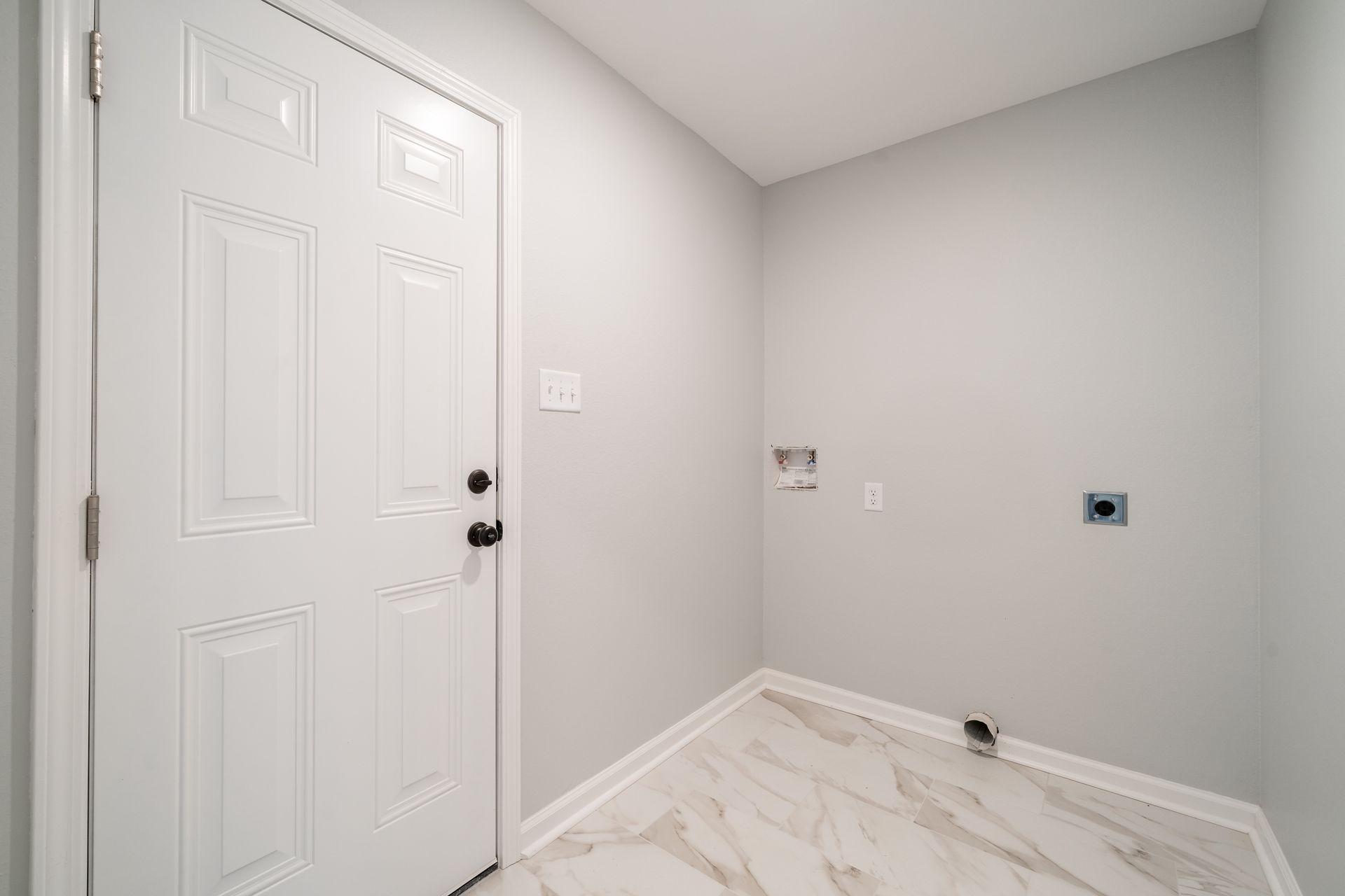 Empty laundry room with white door, gray walls, and light-colored tile floor. Electrical outlets visible.