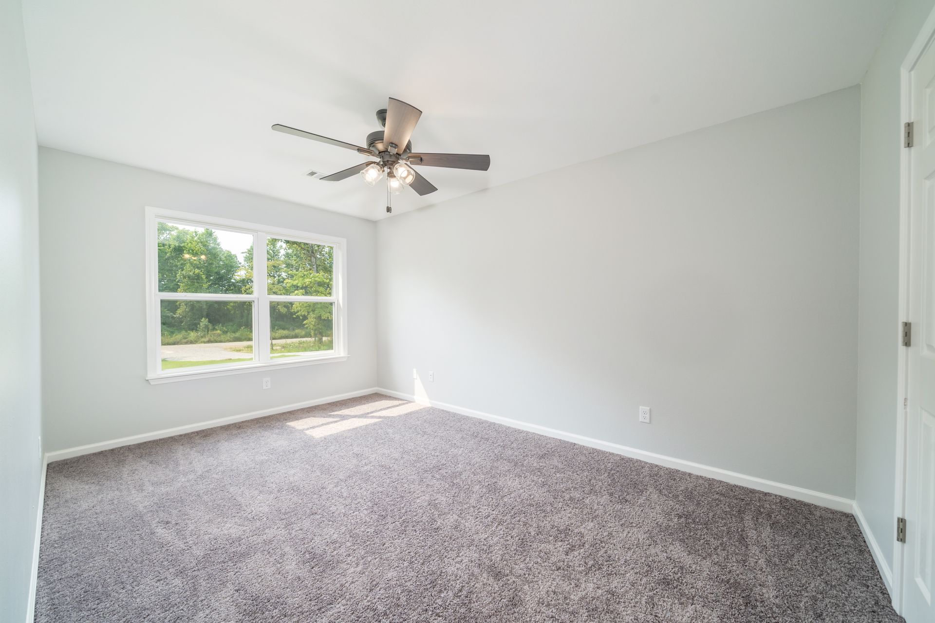 Empty bedroom with gray walls and carpet; window, ceiling fan, and closed door.