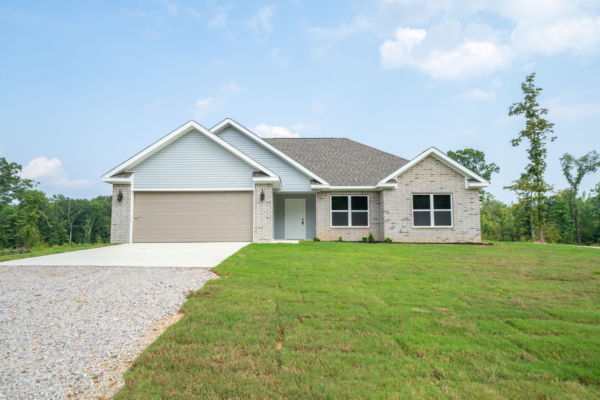 Single-story house with a brick facade, gray roof, and garage, on a grassy lot under a blue sky.