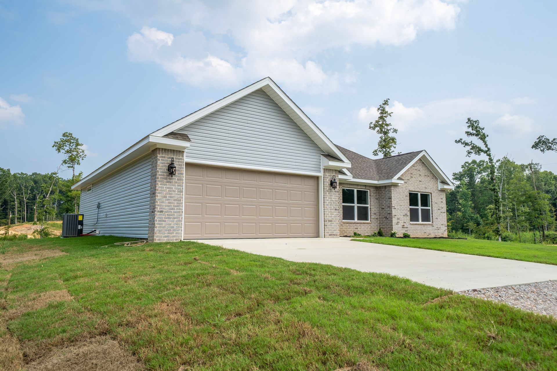 A new single-story house with a light-colored brick and tan garage door on a grassy hill under a blue sky.