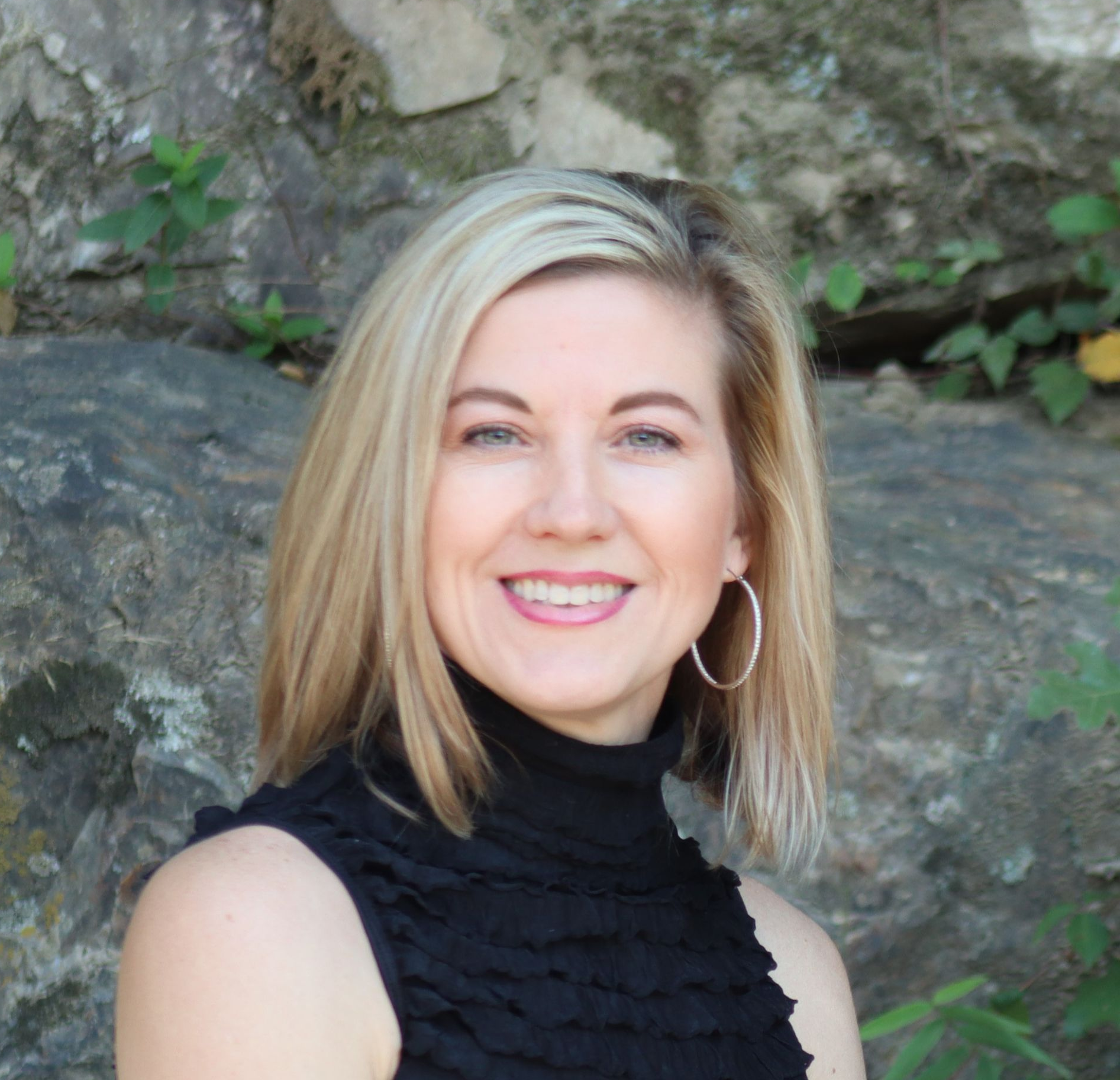Blonde woman smiling, wearing a black top and silver hoop earrings, in front of a stone wall.