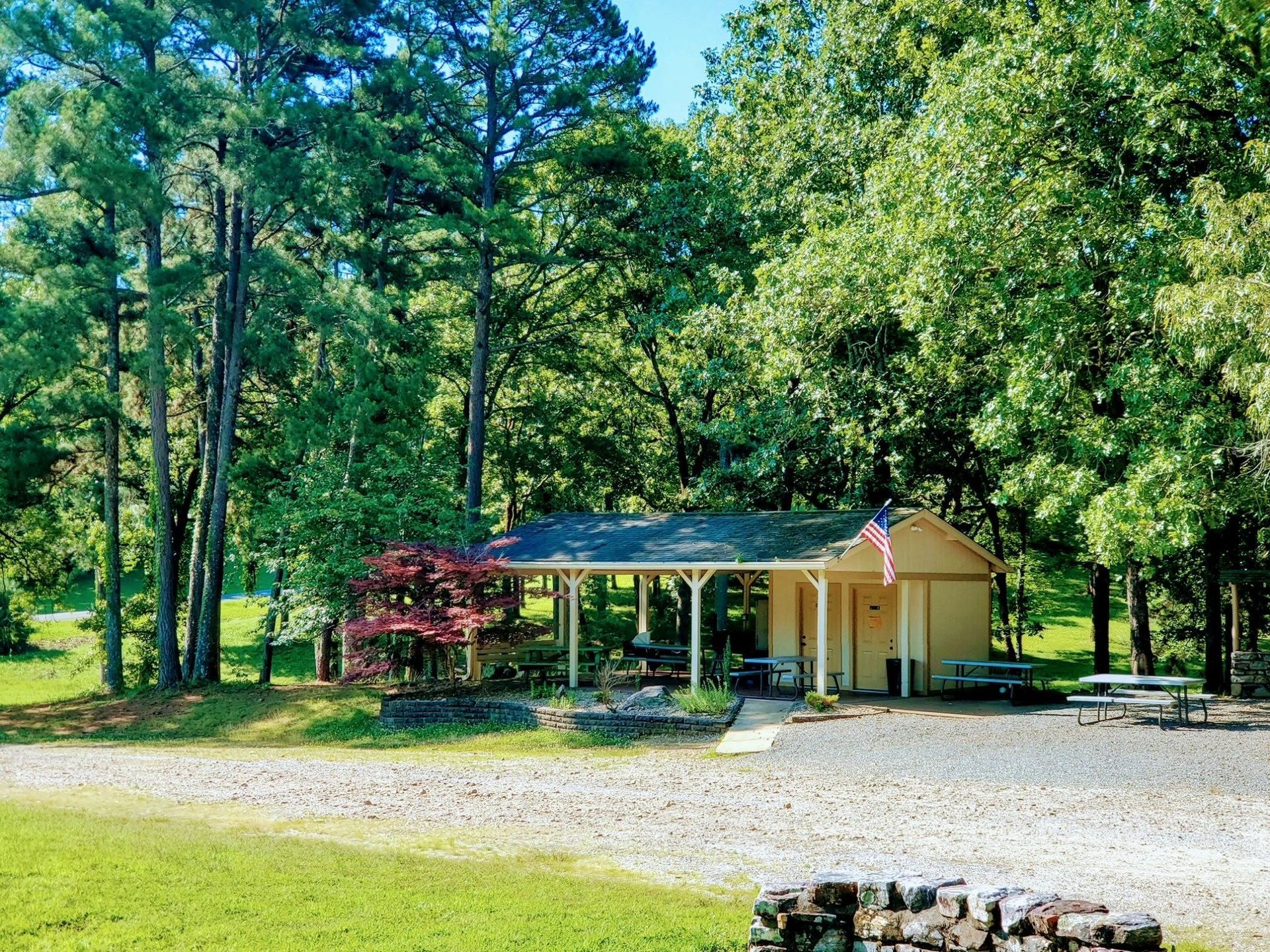 Small, beige building with porch, American flag, surrounded by trees. Gravel drive, green grass.