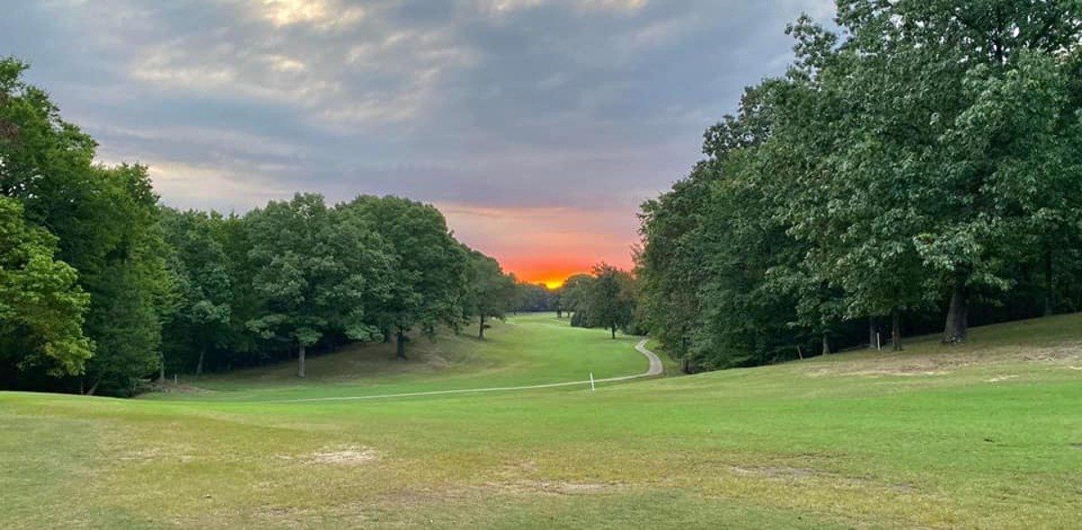 Sunset over a green golf course, framed by trees. Cloudy sky.