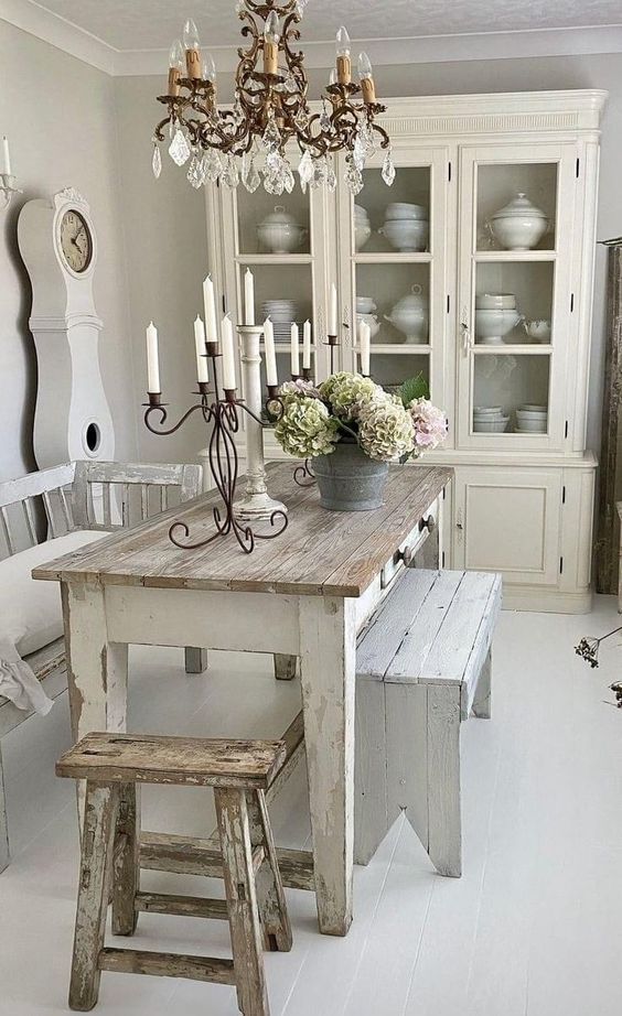 A dining room with distressed white furniture, chandelier, and cabinet filled with china.