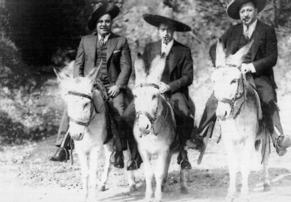 Three men in suits and hats ride donkeys on a dirt path.