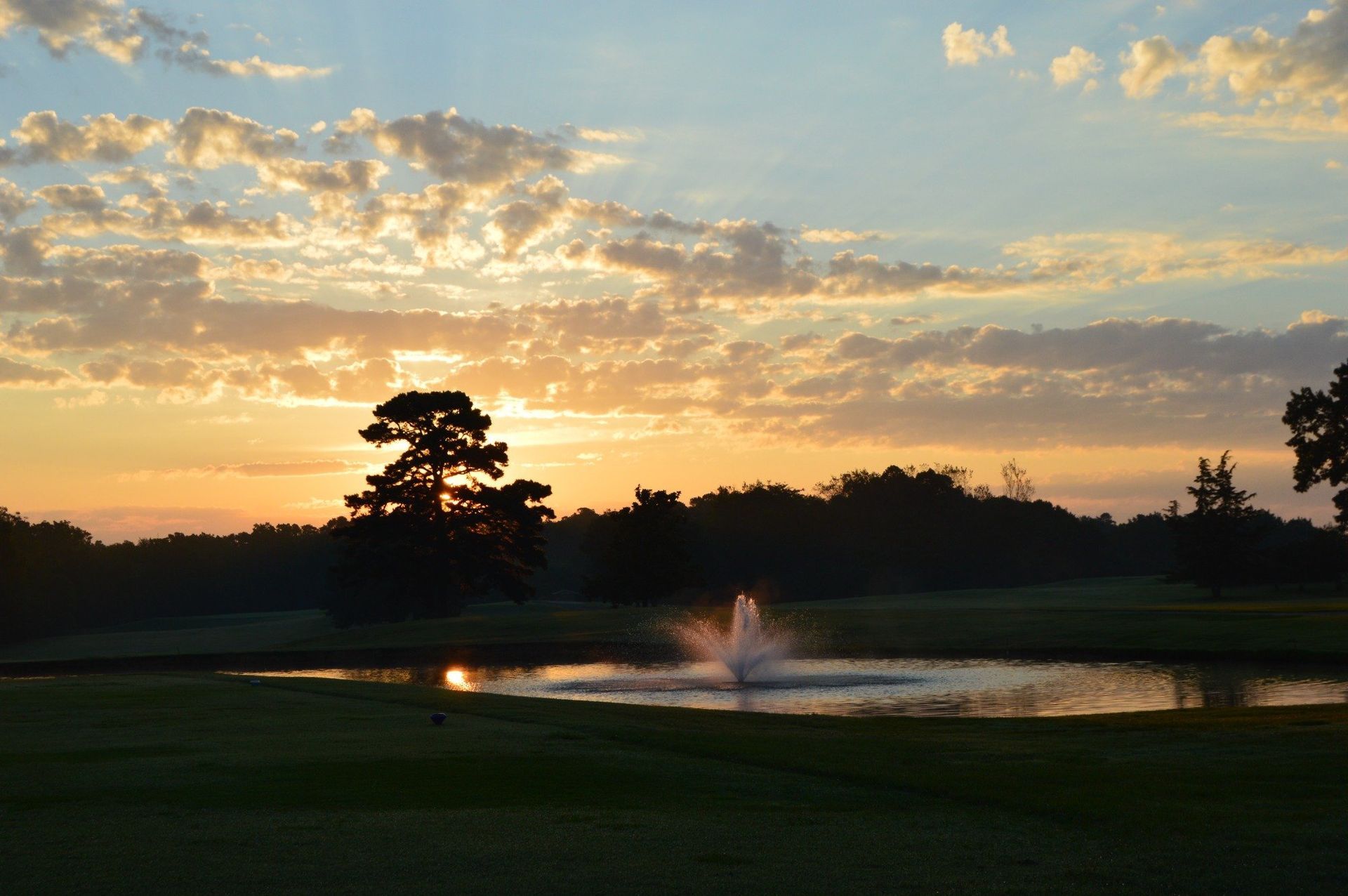 Sunset over a golf course with a pond and fountain, silhouetting trees and illuminating the sky with golden light.
