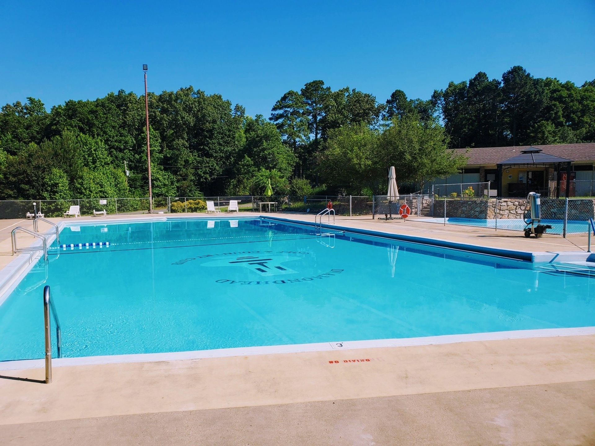 Outdoor swimming pool with blue water, concrete deck, and trees under a clear sky.