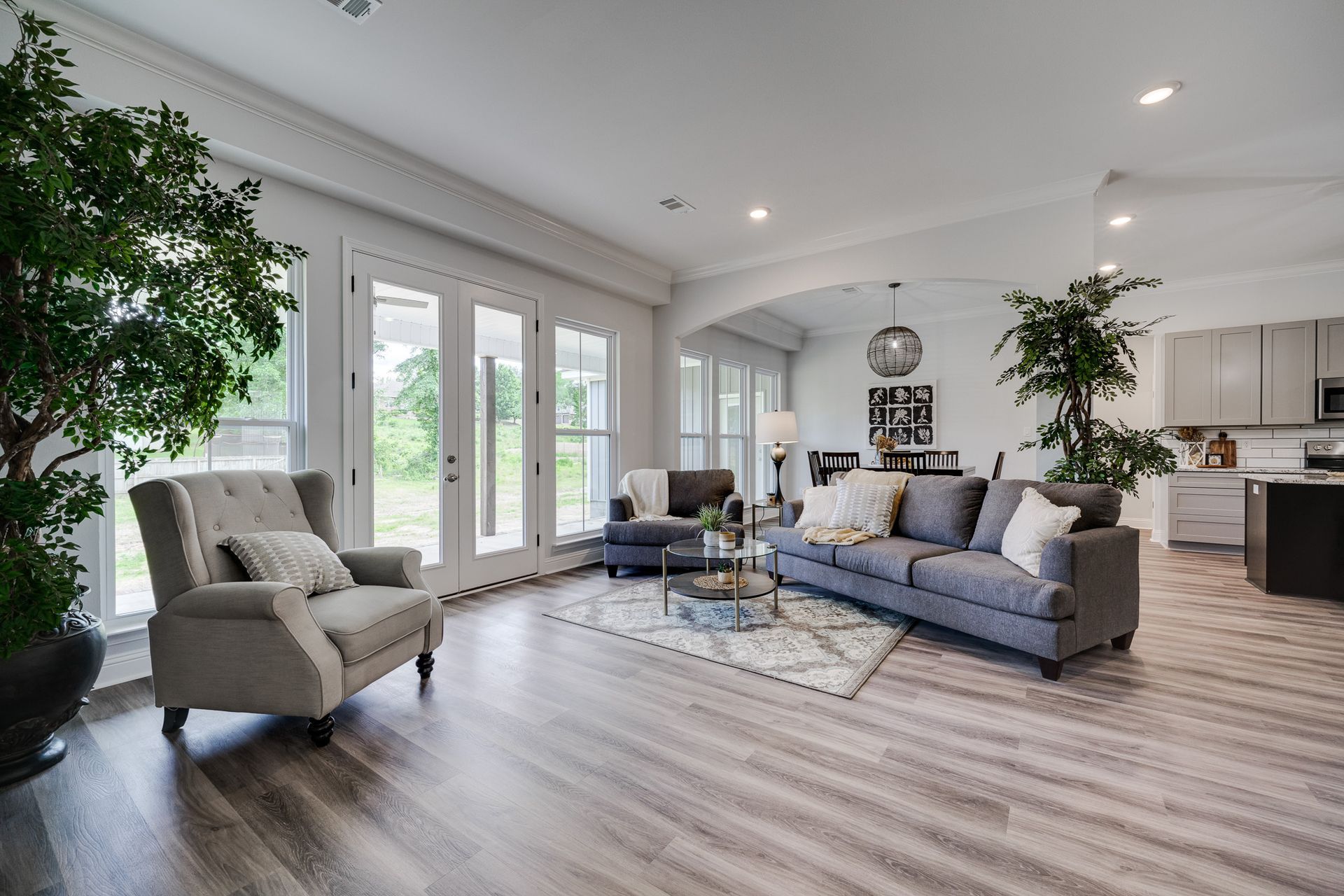 Living room with gray sofa, armchair, and french doors, with hardwood floors and a view of trees.