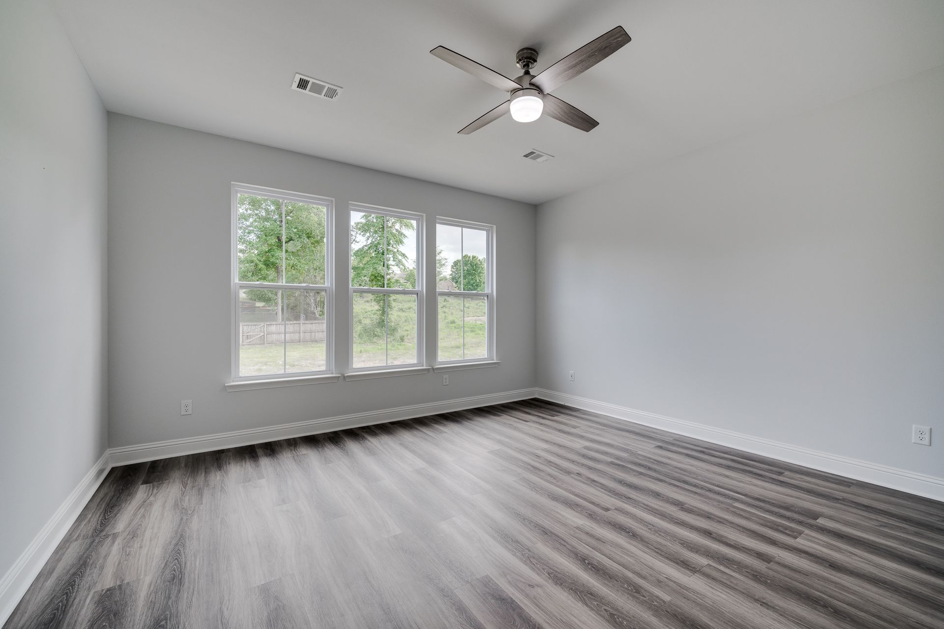 Empty bedroom with grey walls, three windows, wood-look flooring, and a ceiling fan.