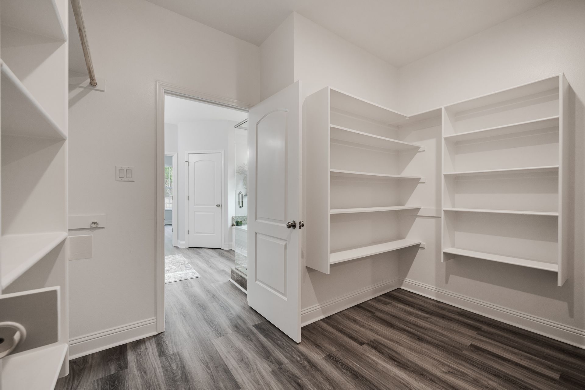White pantry with shelves, open door to a bathroom with a white door. Dark wood-look floors.