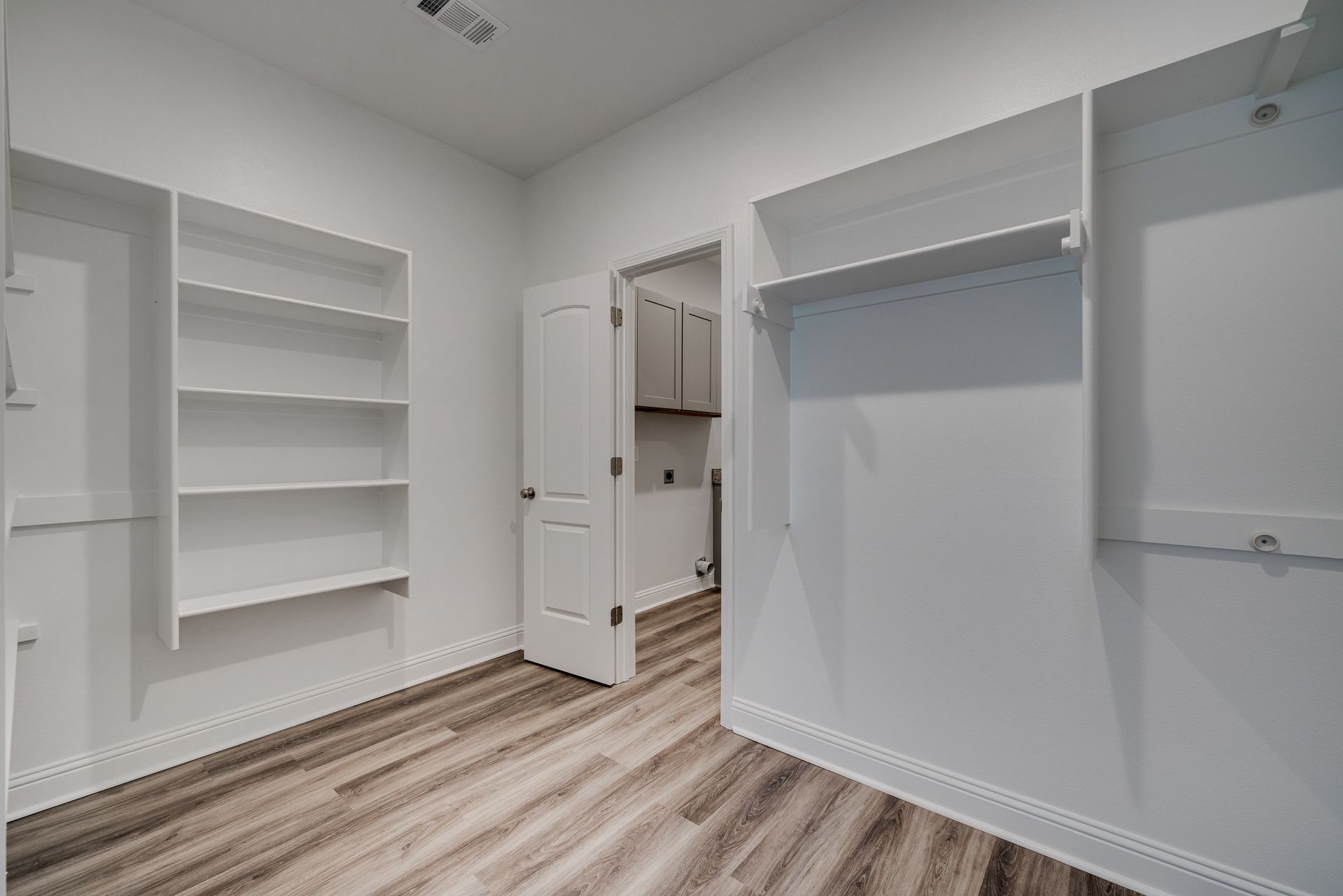 Empty white walk-in closet with shelves, wood-look floor, and an open door to another room.