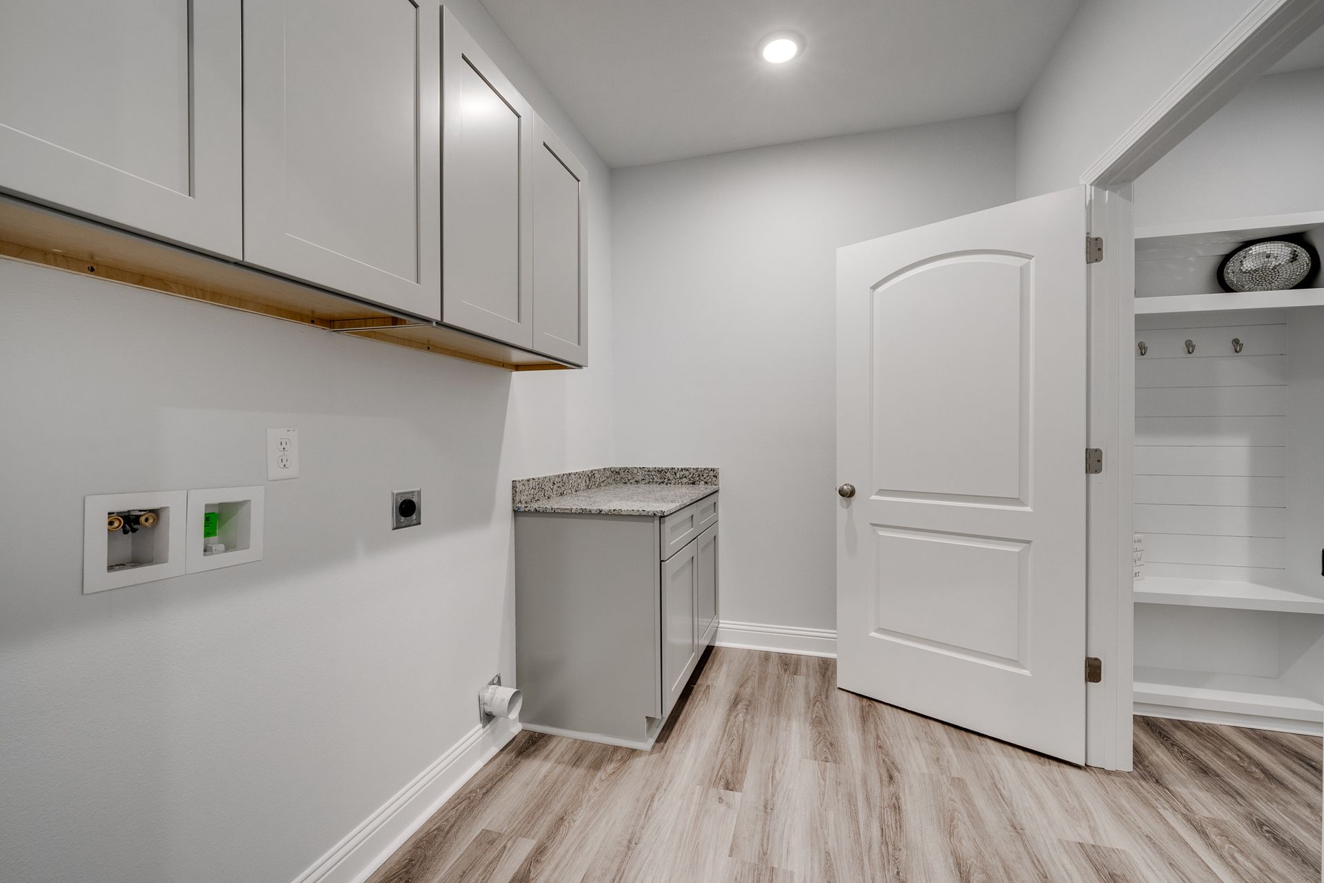 Laundry room with gray cabinets, countertop, white door, and shelving. Wooden floor.