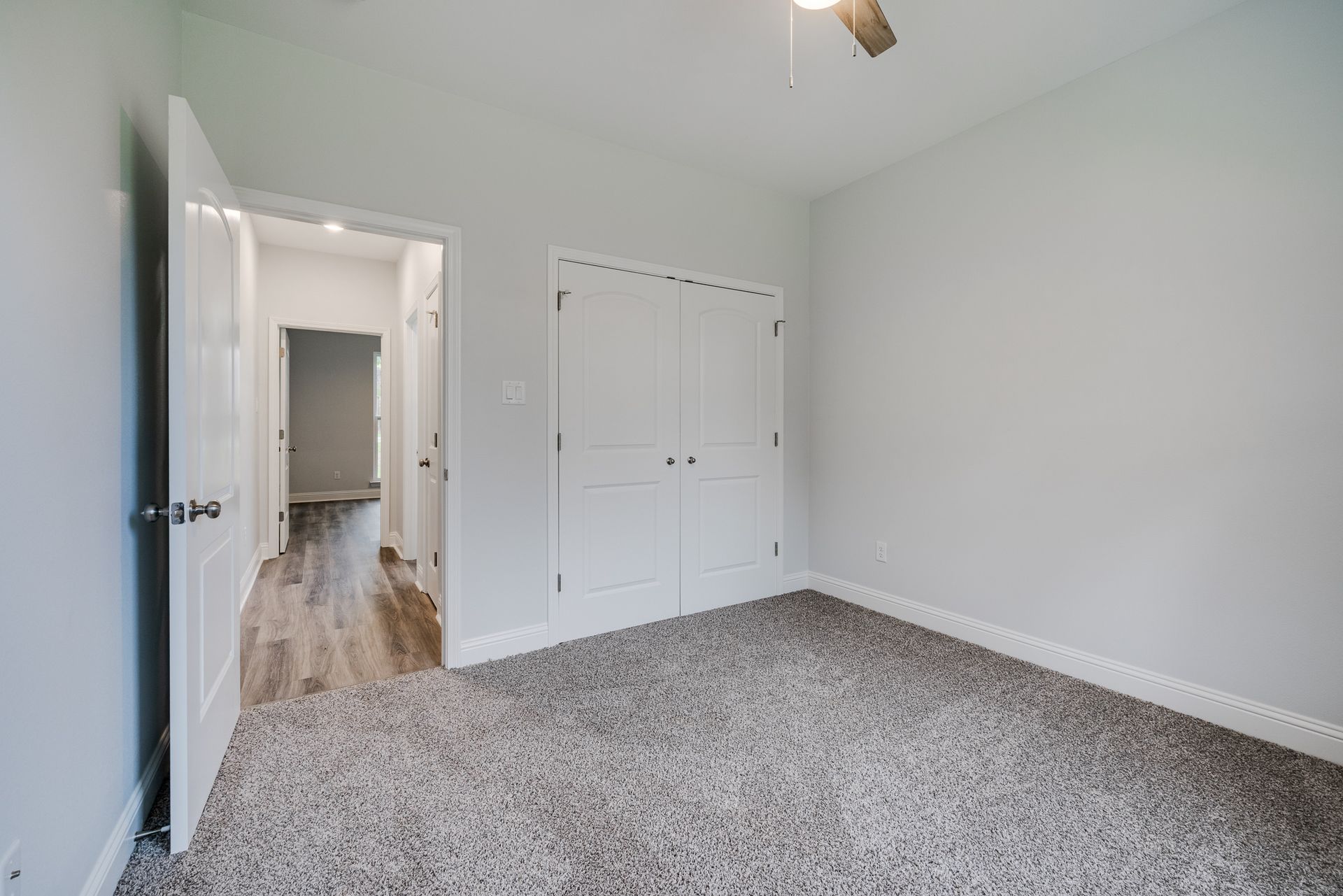Empty bedroom with gray carpet, white closet doors, and a hallway visible through an open doorway.