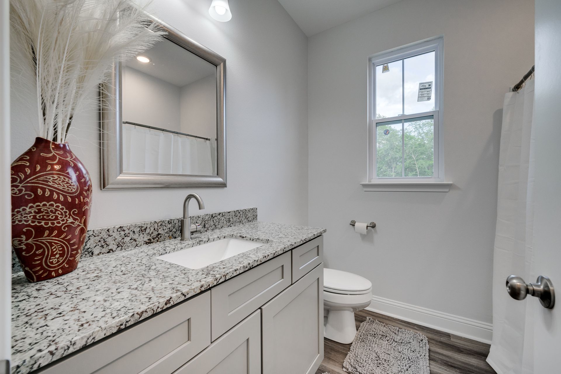 Bathroom with gray vanity, speckled countertop, white toilet, and window. Red vase with white flowers.