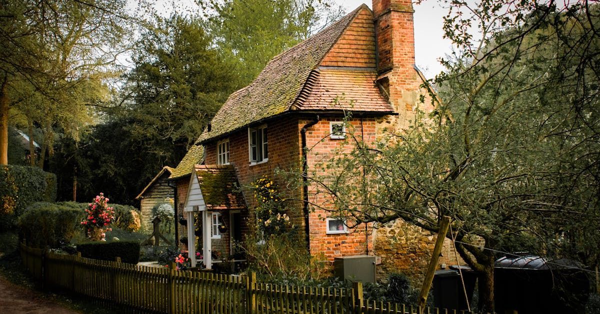 Cottage with a thatched roof, brick facade, and a small picket fence, nestled among trees.