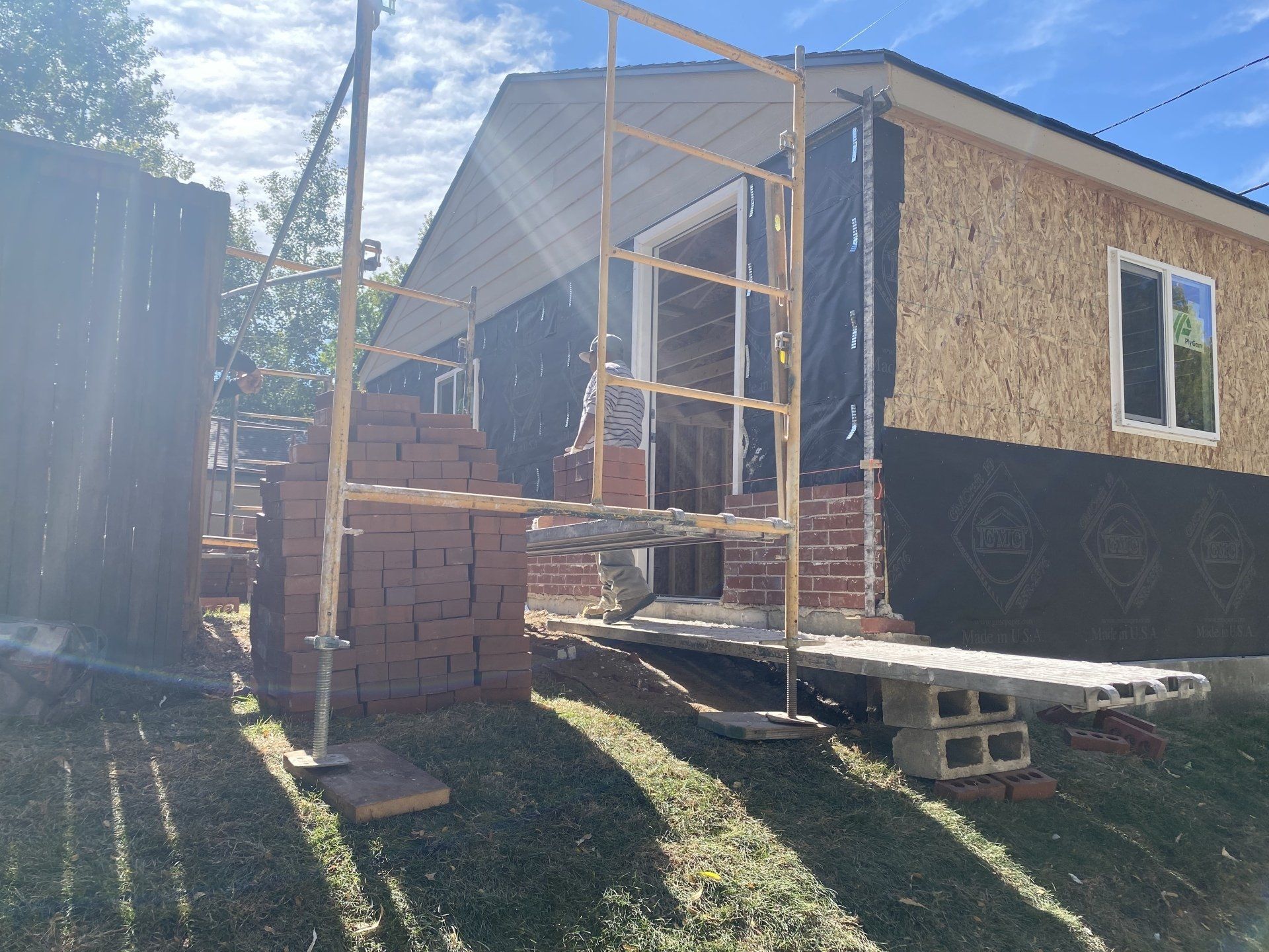 Bricklaying in progress on a house exterior. Scaffolding, bricks, black wrap visible. Green grass, sunny day.