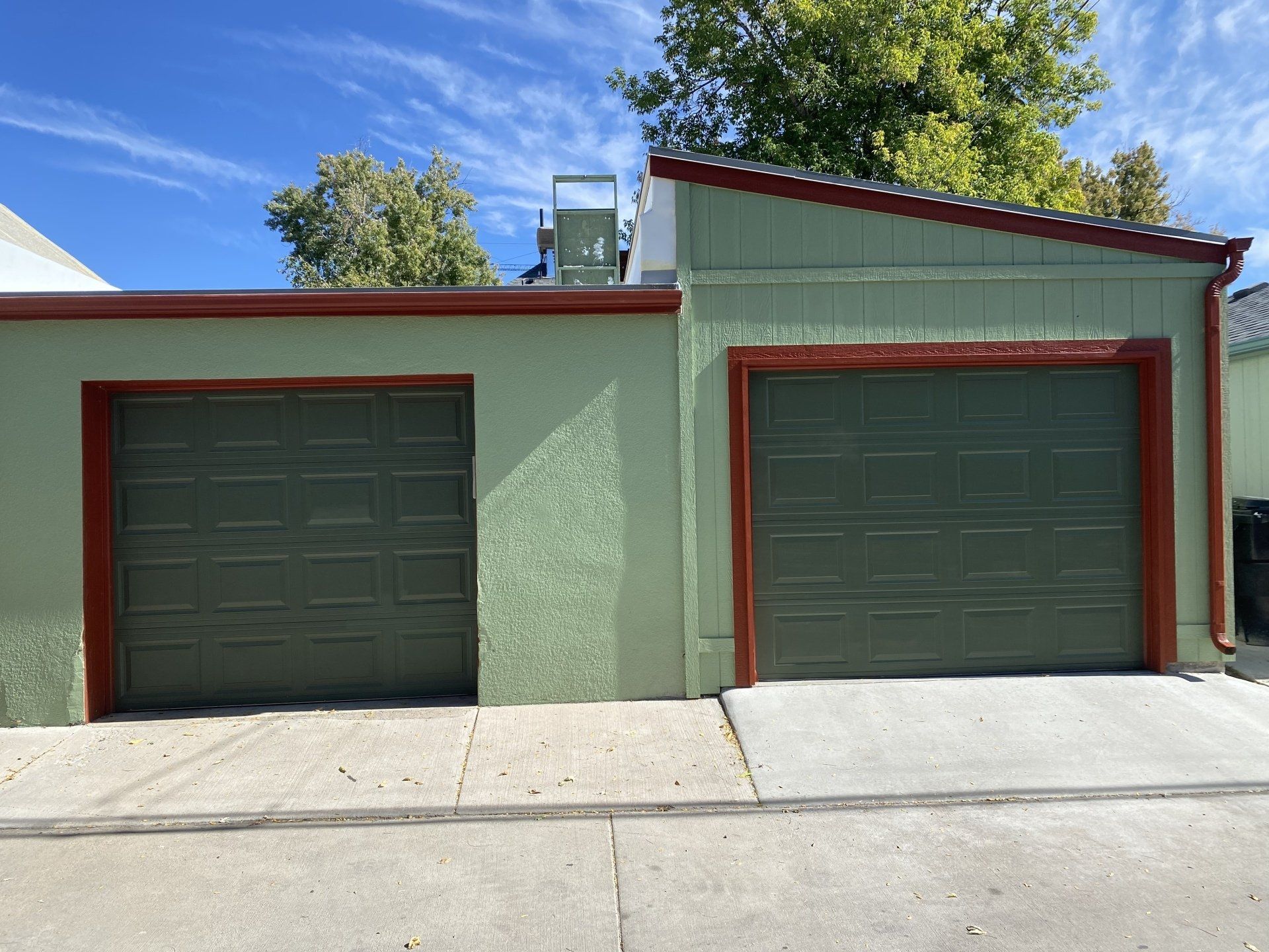 Two green garage doors with brown trim against a stucco building, under a blue sky.