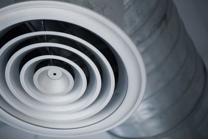 A circular white air diffuser vent mounted on a metallic industrial ceiling duct.
