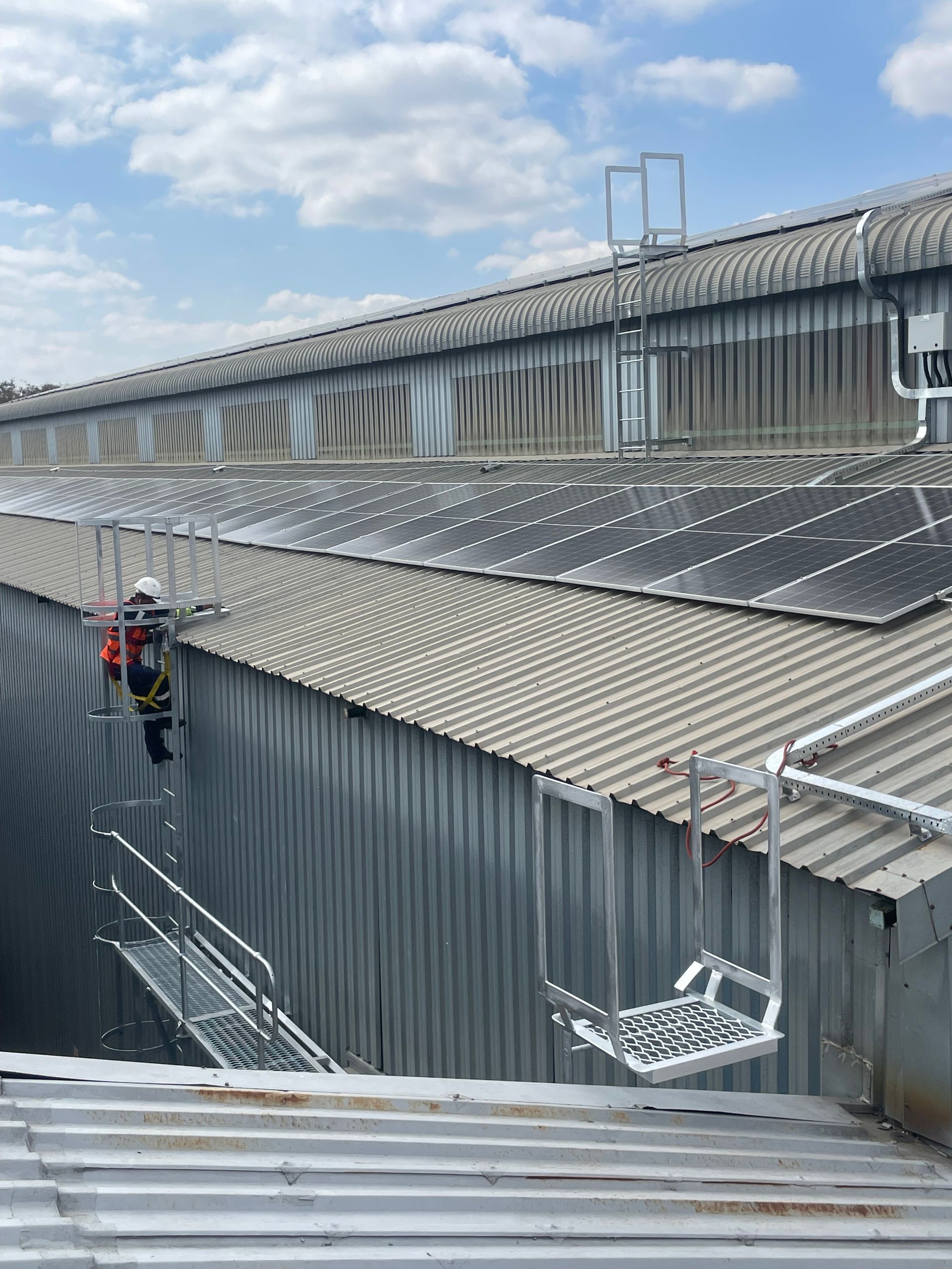 Person on metal ladder accessing building roof. Cloudy sky. Grey metal building with solar panels.