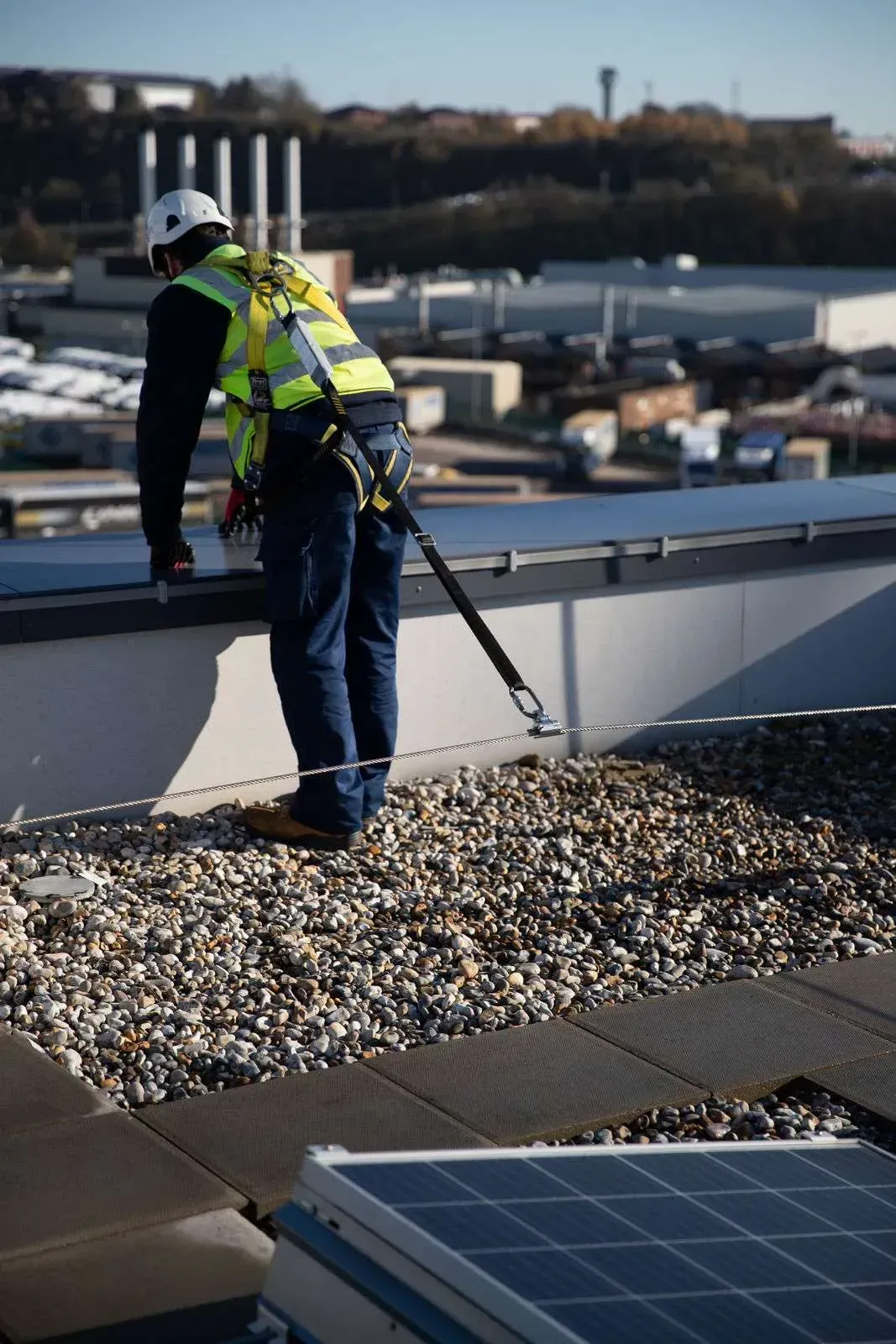 Person in safety harness on a rooftop, looking out. Wearing a hard hat and high-visibility vest. Blue sky.