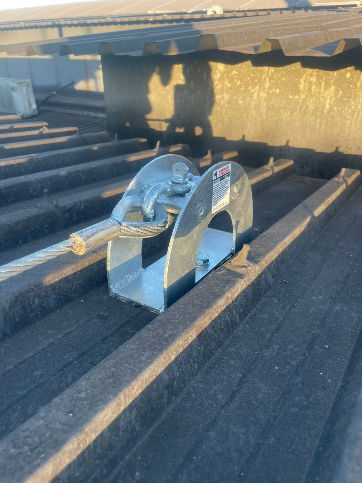 Silver fall arrest system secured to a dark corrugated metal roof. A metal cable extends from the device.