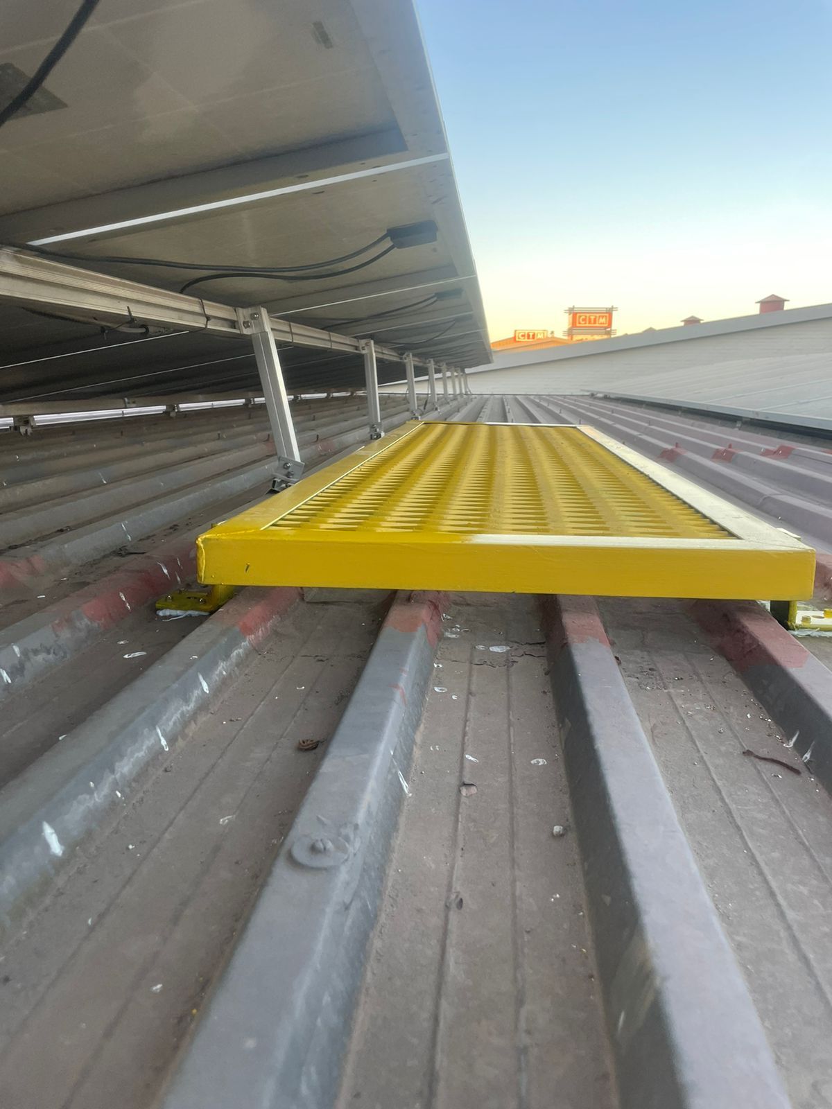 Yellow rooftop walkway installed on a corrugated metal roof, beneath solar panels.
