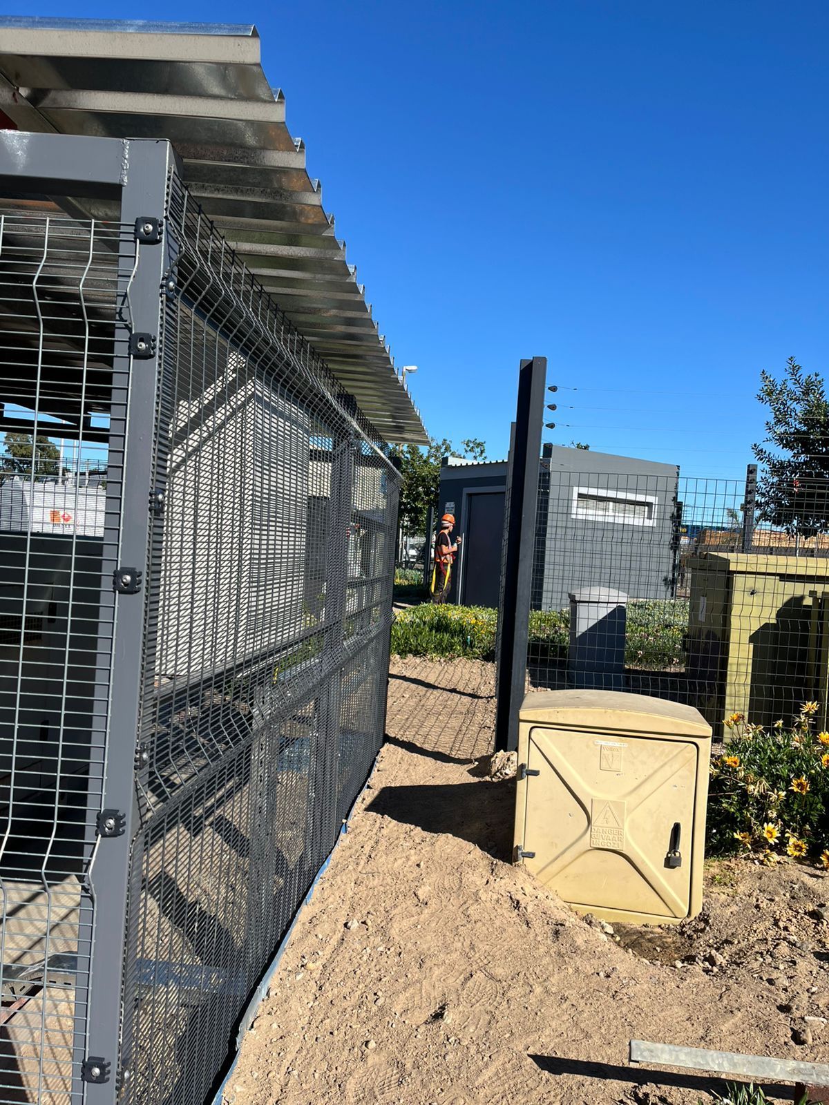 Gray metal fence and shed with gravel ground under a blue sky.