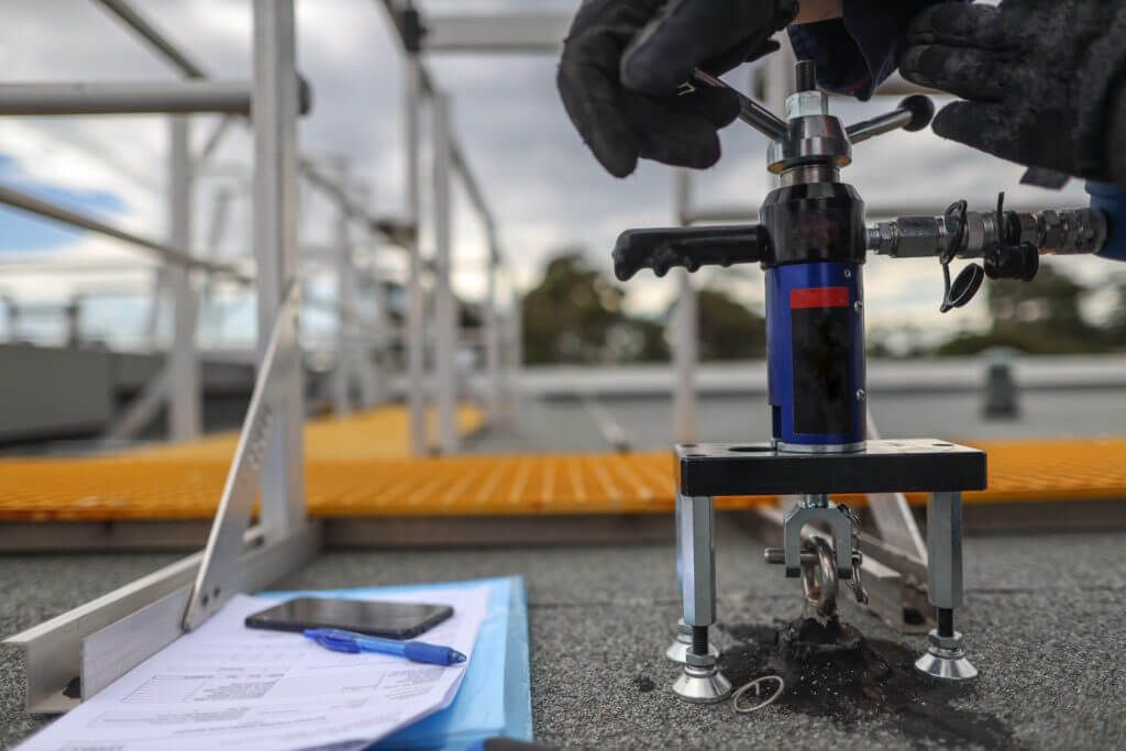 Gloved hands operate a testing device on a rooftop. A clipboard and scaffold are in the background.