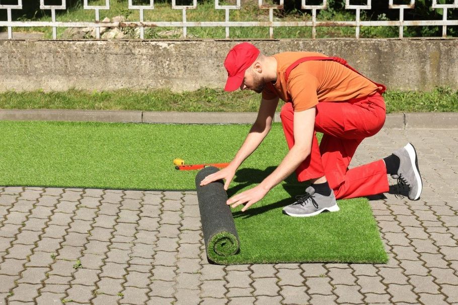 Worker in red kneels, rolling out green turf on a paved lawn.