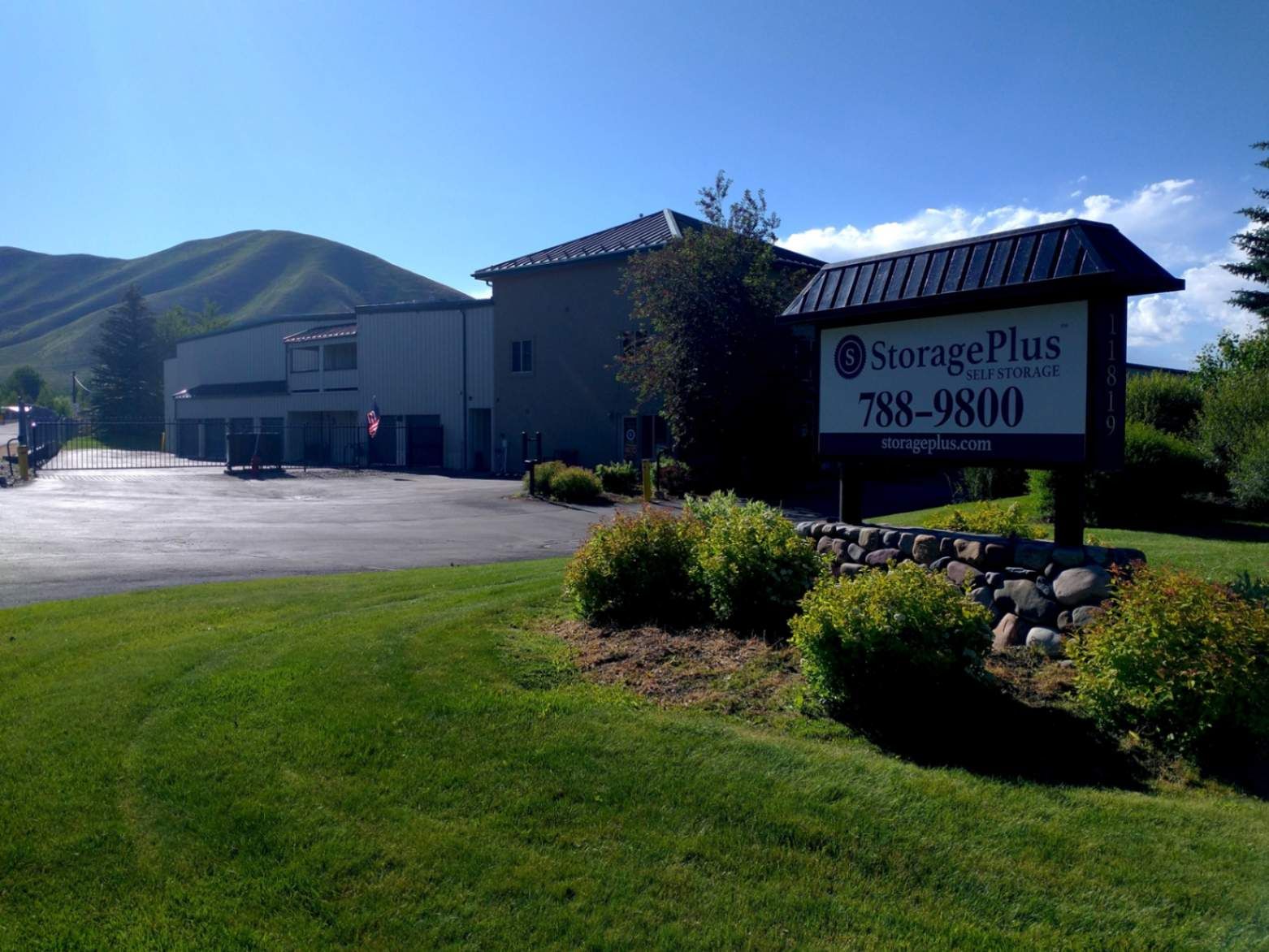 Storage Plus facility with sign, building, and gate on a sunny day. Green grass and hills in the background.