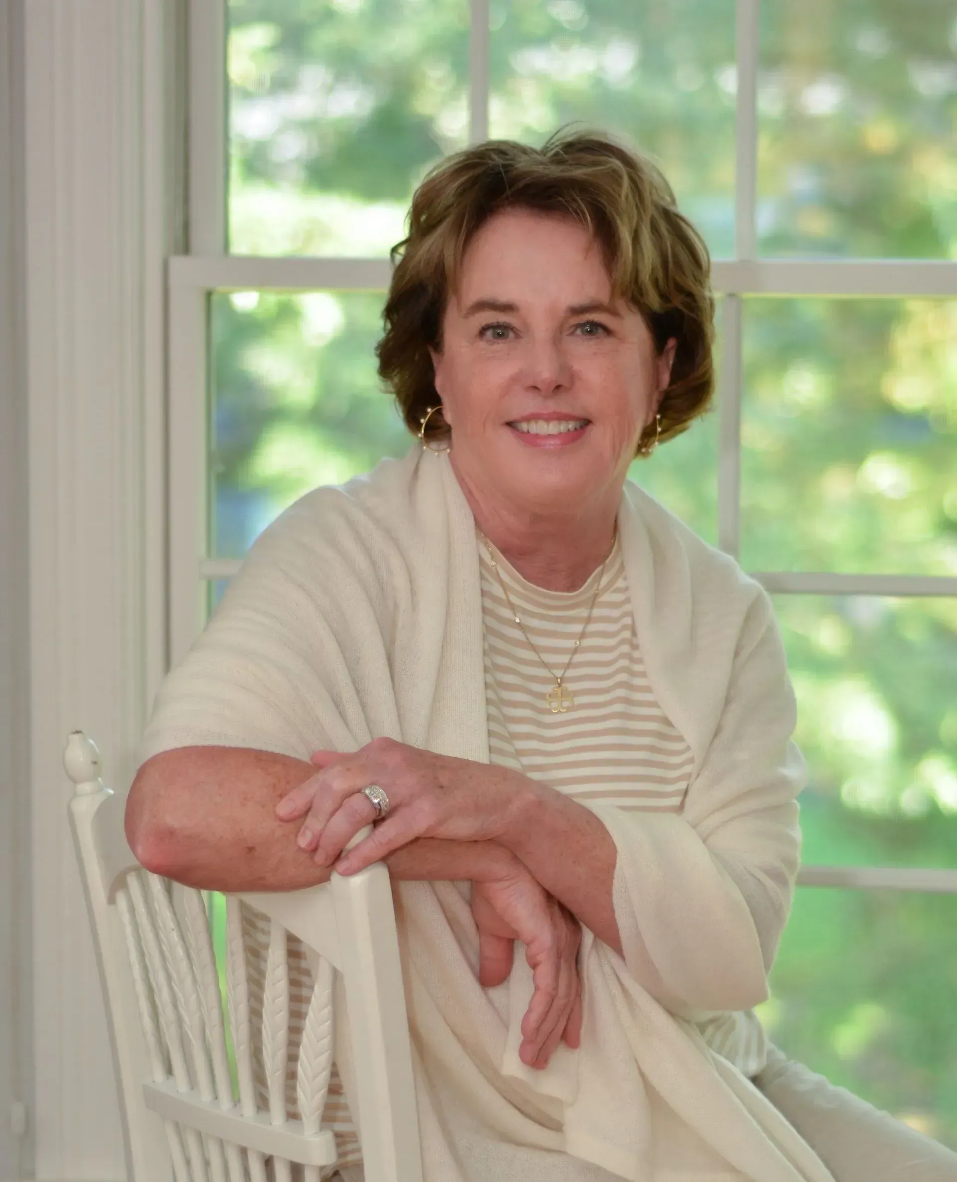 A woman is sitting in a white chair in front of a window.