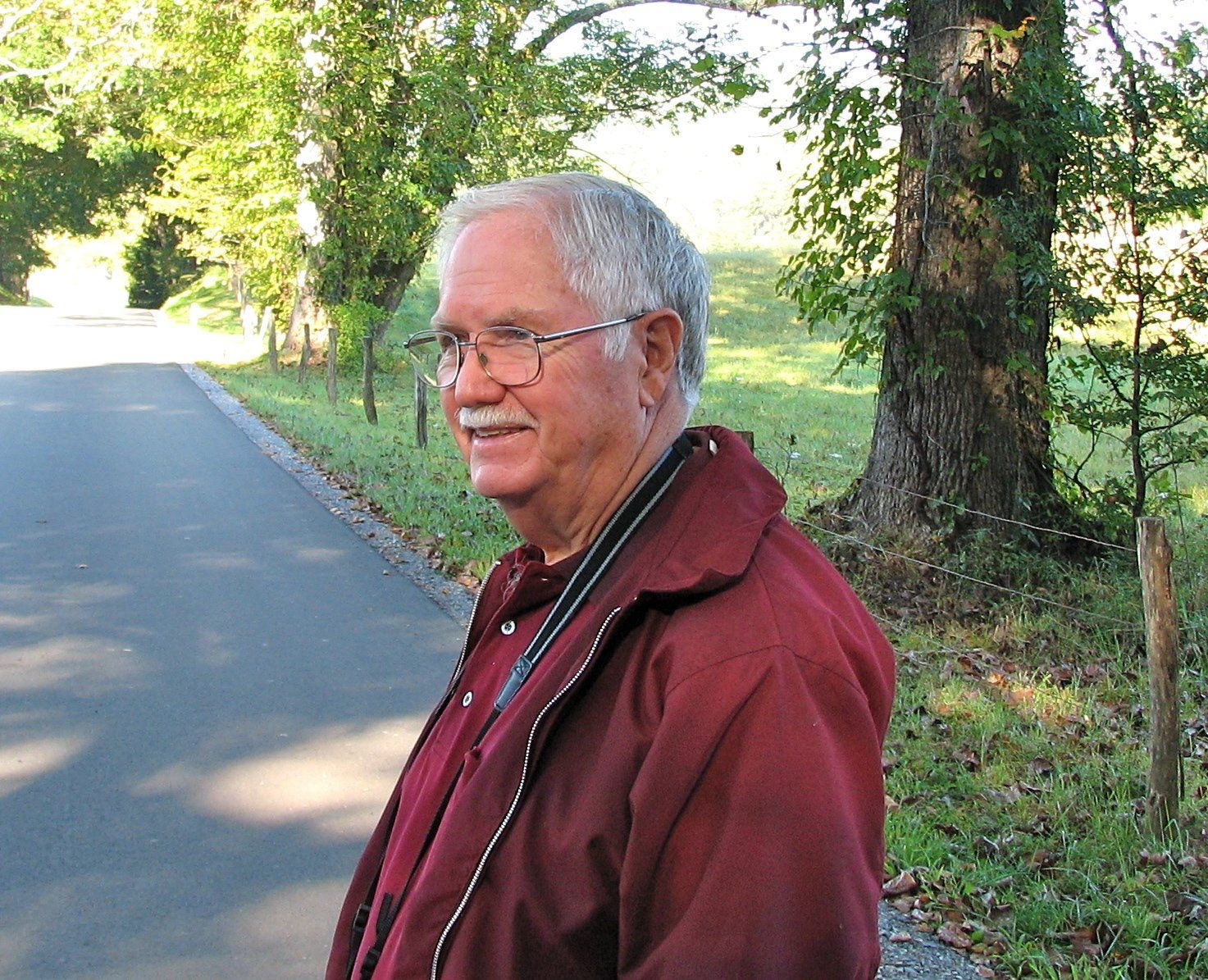 A man wearing glasses and a red jacket stands on the side of a road