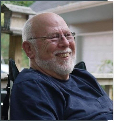 A man with glasses and a beard is smiling while sitting in a wheelchair