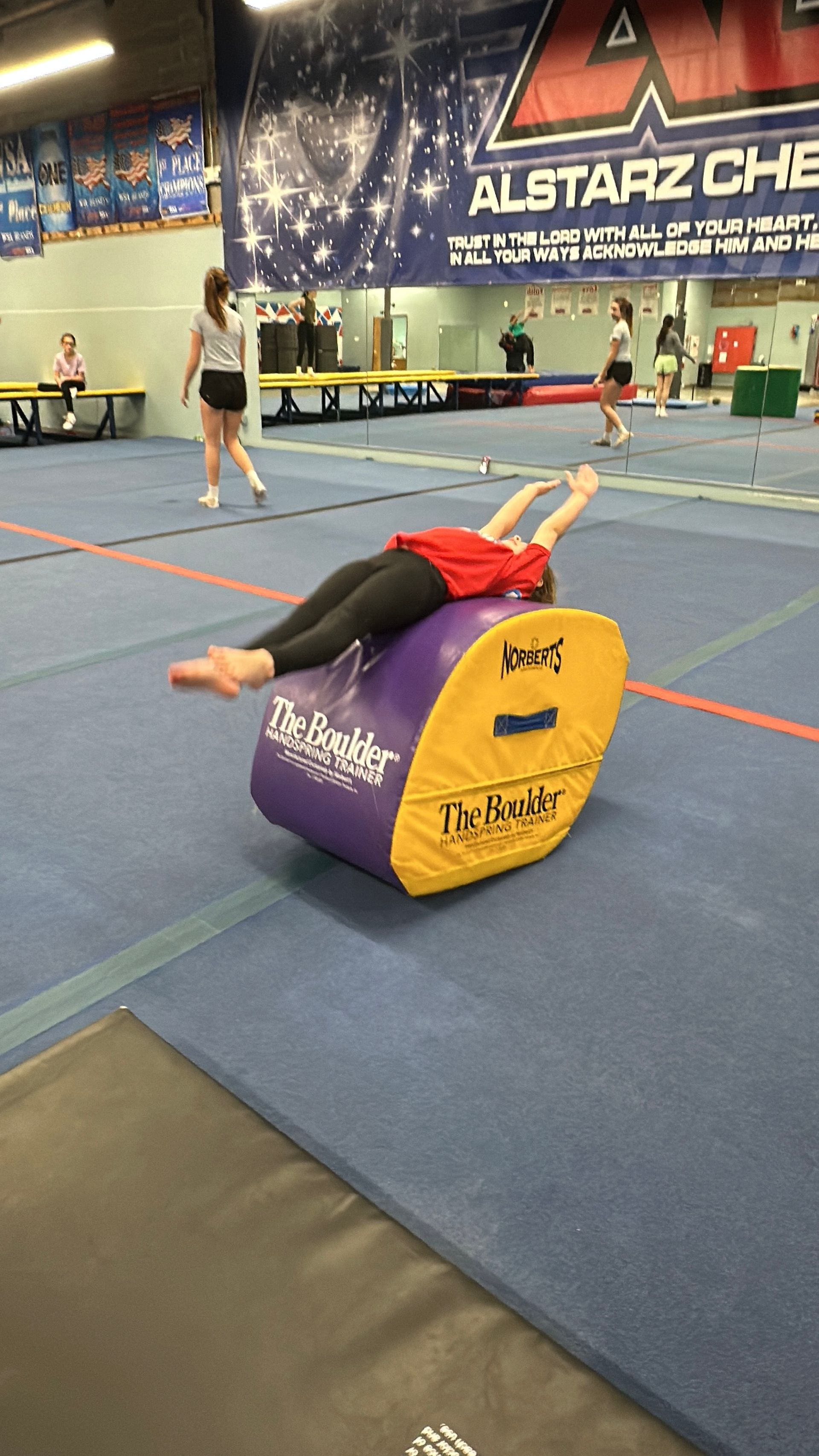 A woman is laying on top of a purple and yellow barrel in a gym.