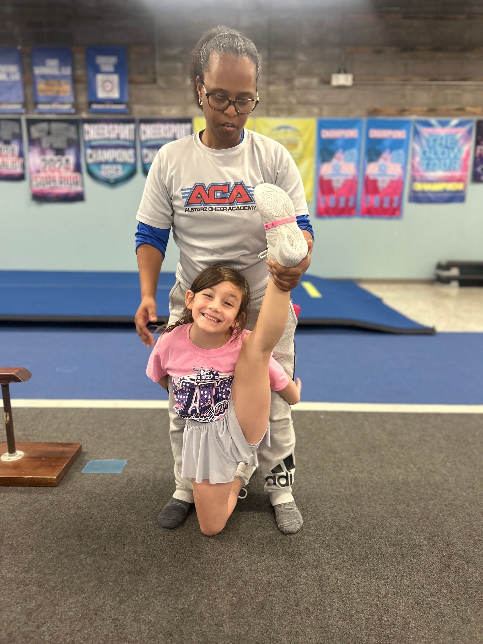 A woman is helping a little girl do a split in a gym.