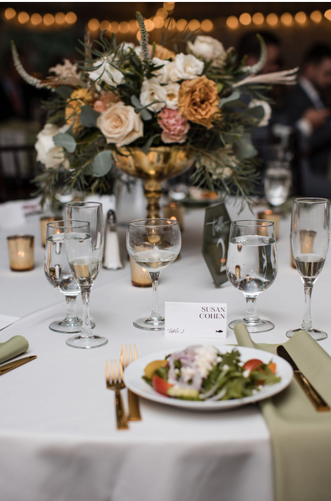 A table set for a wedding reception with plates of food and wine glasses.
