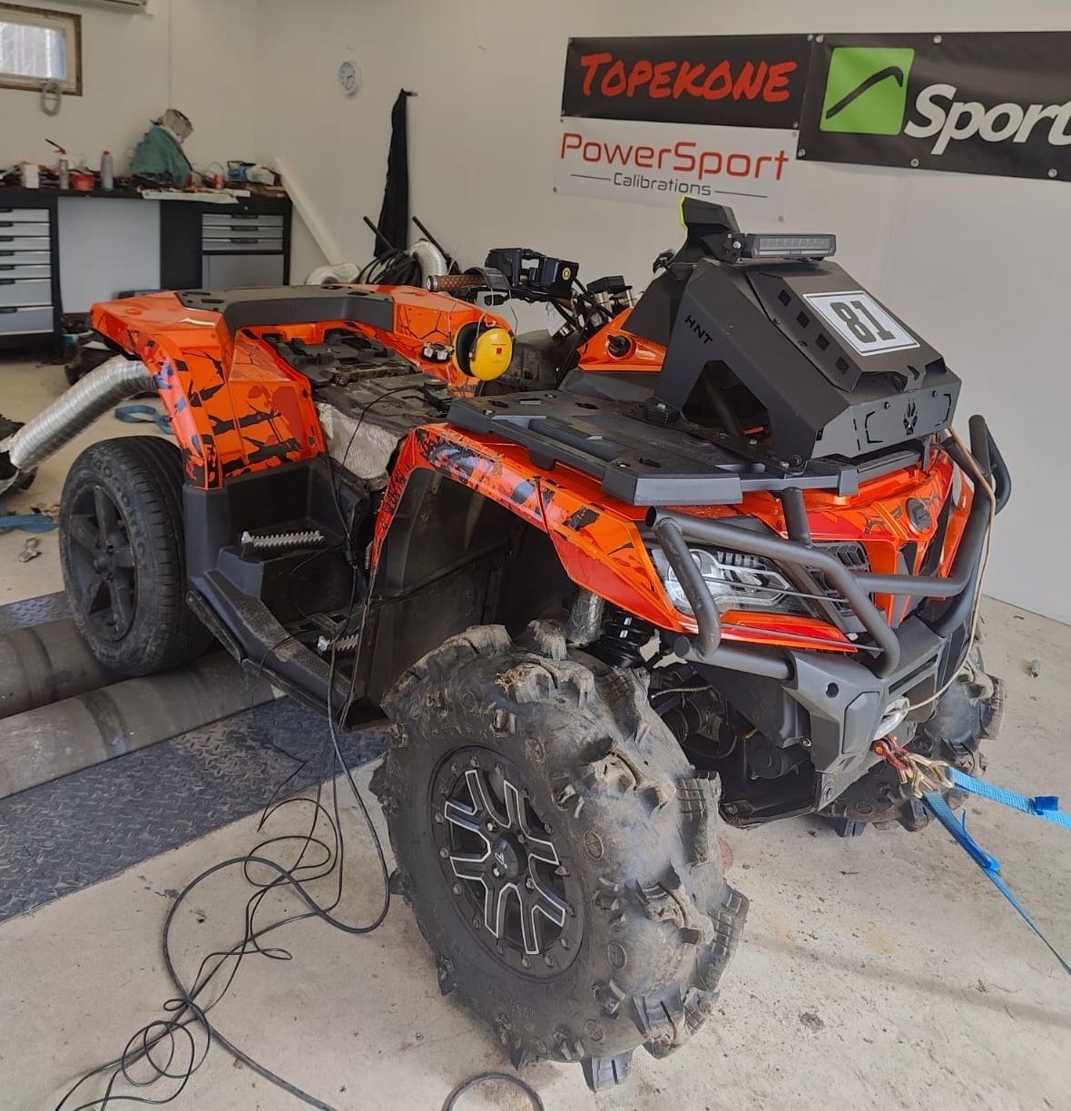Orange-and-black ATV in a garage, muddy tires, with tools and a PowerSport banner on the wall