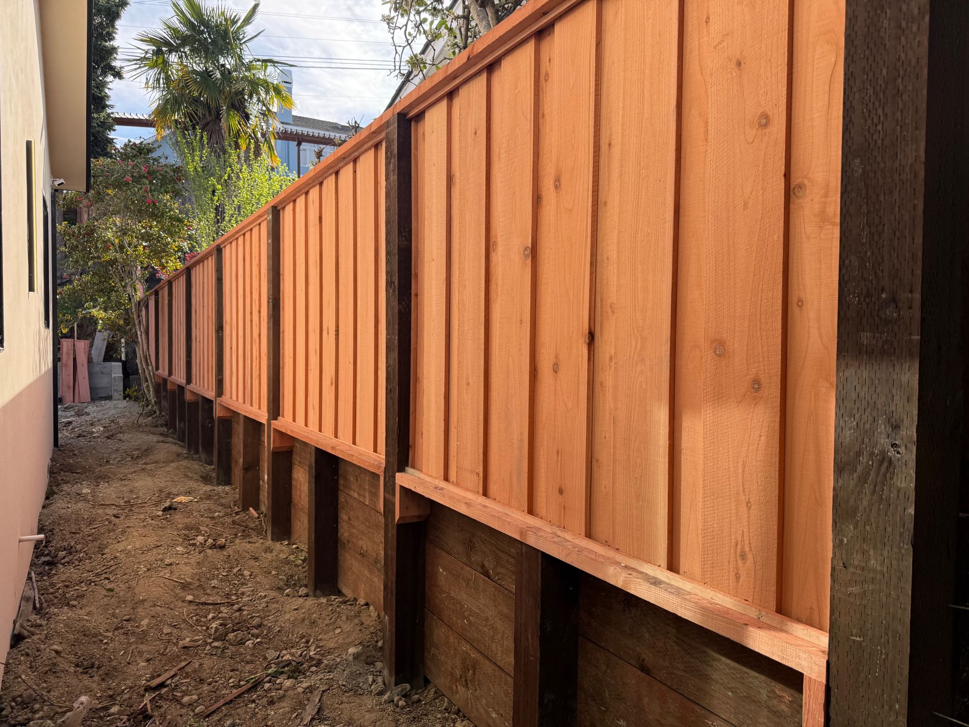 Wooden fence with vertical planks, light brown, along a dirt path next to a building.