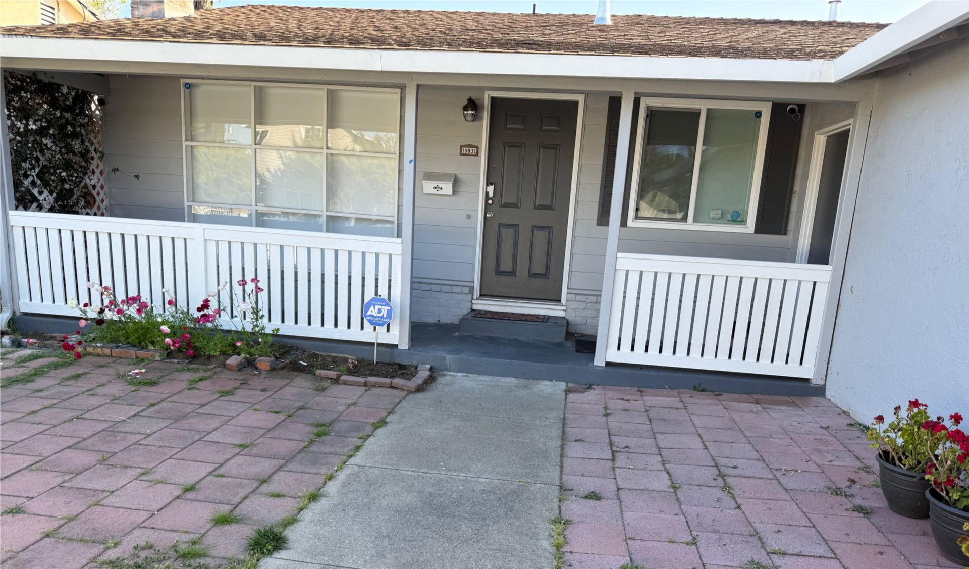 Gray house with white railing and red brick walkway. Dark door, windows, and potted flowers.
